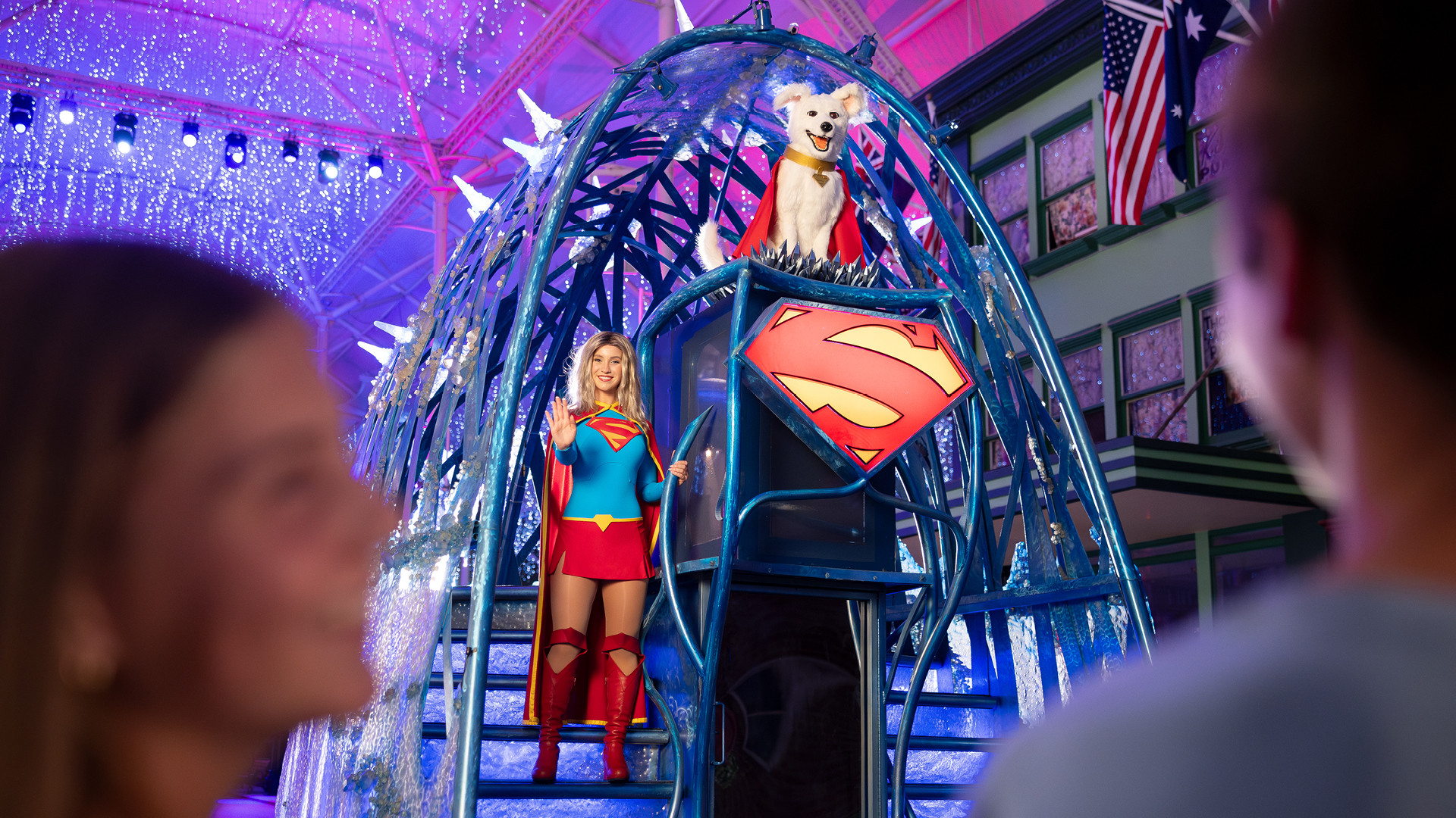 A woman dressed as Supergirl stands on a decorated float with a dog wearing a cape above her, under colorful lights, while two people watch in the foreground.