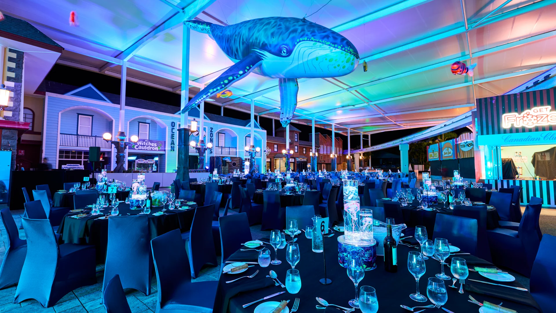 A large event space with round tables set for dinner under blue and purple lighting. A giant inflatable whale is suspended from the ceiling. The area is decorated with elegant table settings and surrounded by building facades.