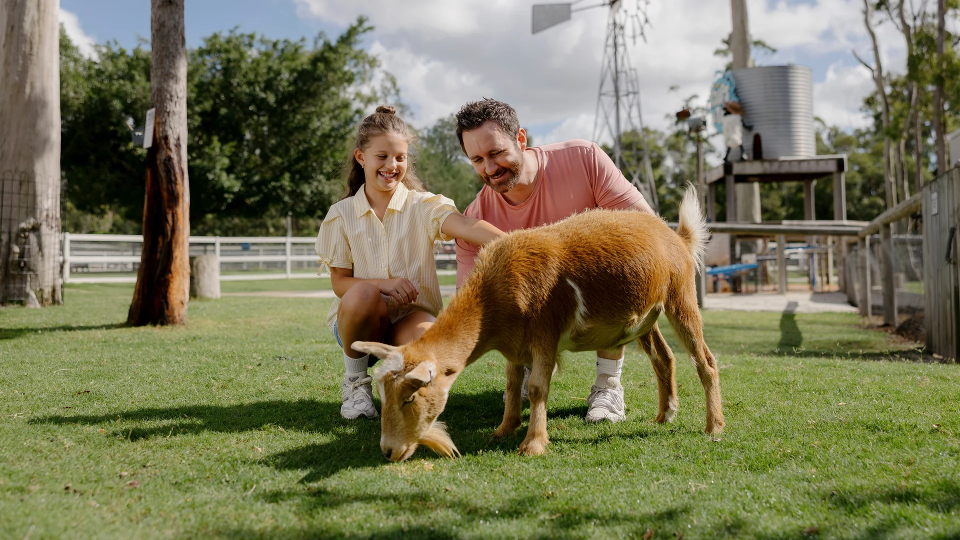 A man and a young girl kneel on grass, smiling as they pet a brown goat that is grazing. Trees, fencing, and a windmill are visible in the background on a sunny day.