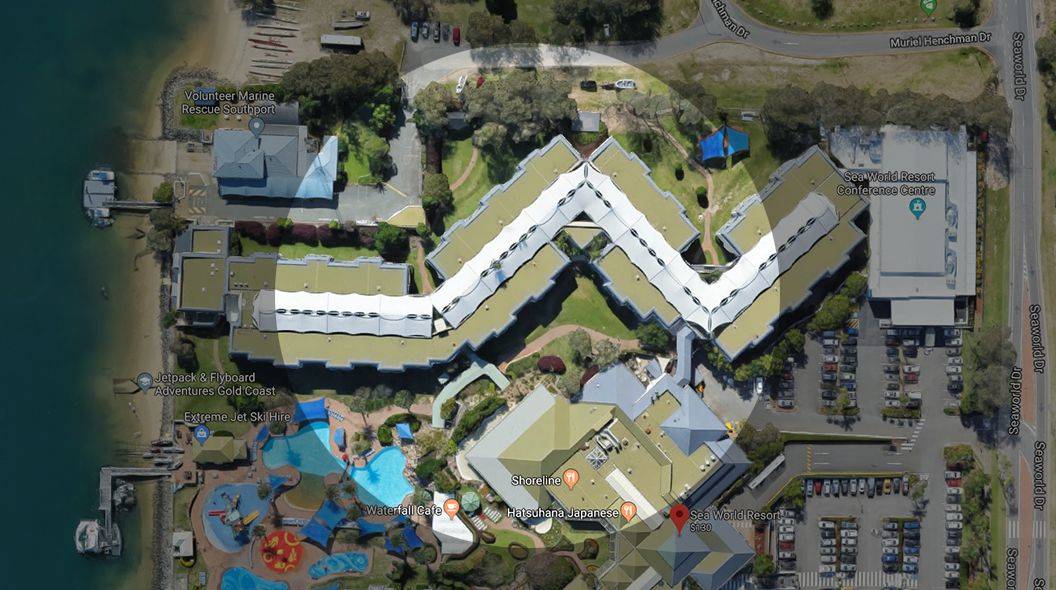 Aerial view of a large building with a zigzag shape near a body of water. Surrounding areas include parking lots, trees, and colorful structures resembling a playground or waterpark.