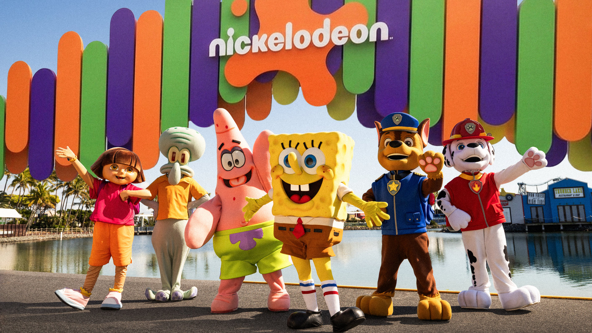 Six Nickelodeon characters, including SpongeBob, Patrick, Squidward, Dora, Chase, and Marshall, pose in front of a colorful Nickelodeon sign and water at an amusement park.