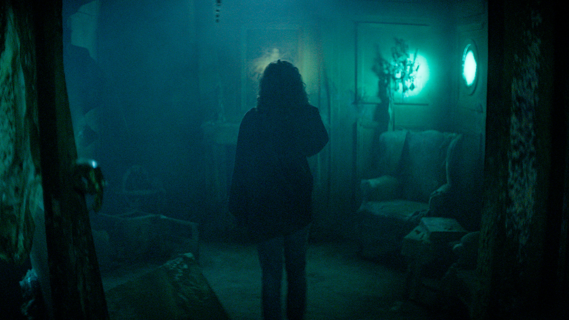 A person with curly hair stands in a dim, foggy room filled with old furniture and eerie greenish-blue lighting, casting a mysterious and haunting atmosphere.