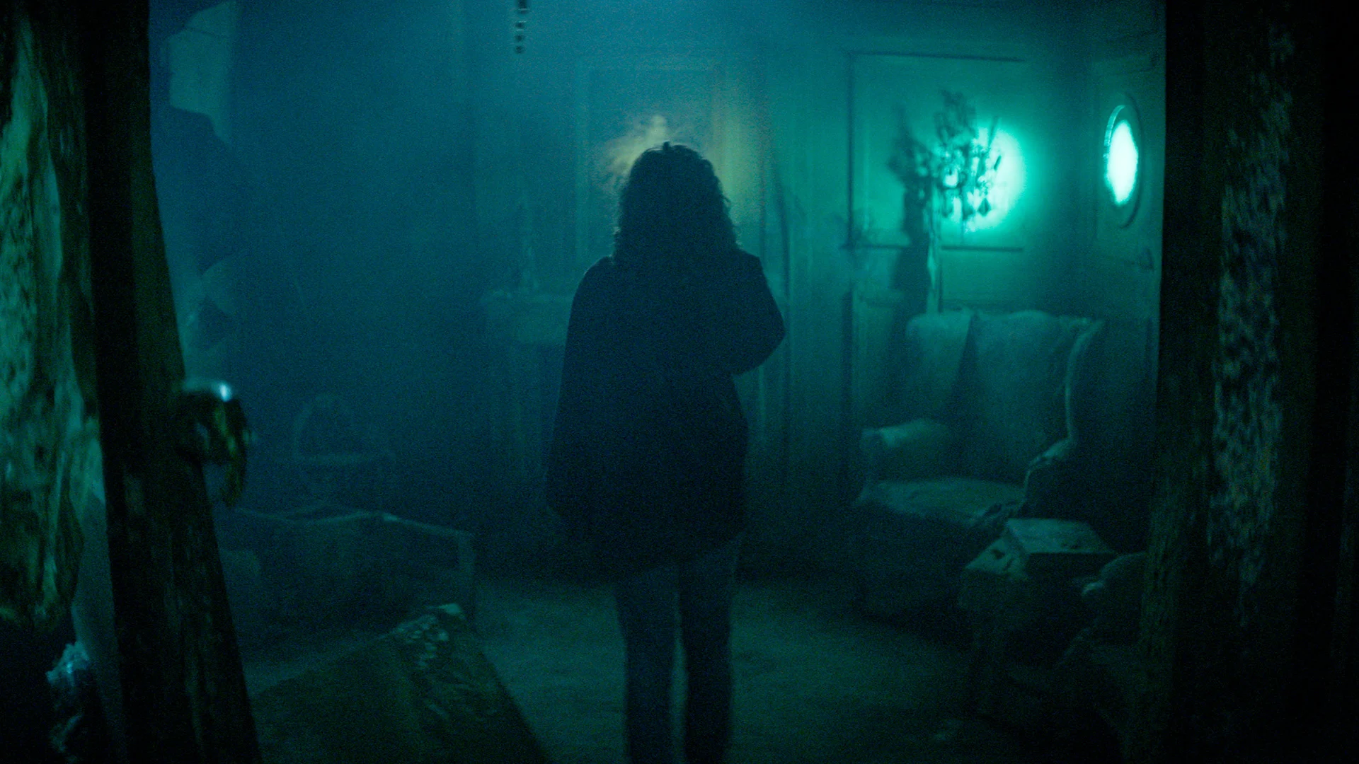 A person with curly hair stands in a dim, foggy room filled with old furniture and eerie greenish-blue lighting, casting a mysterious and haunting atmosphere.