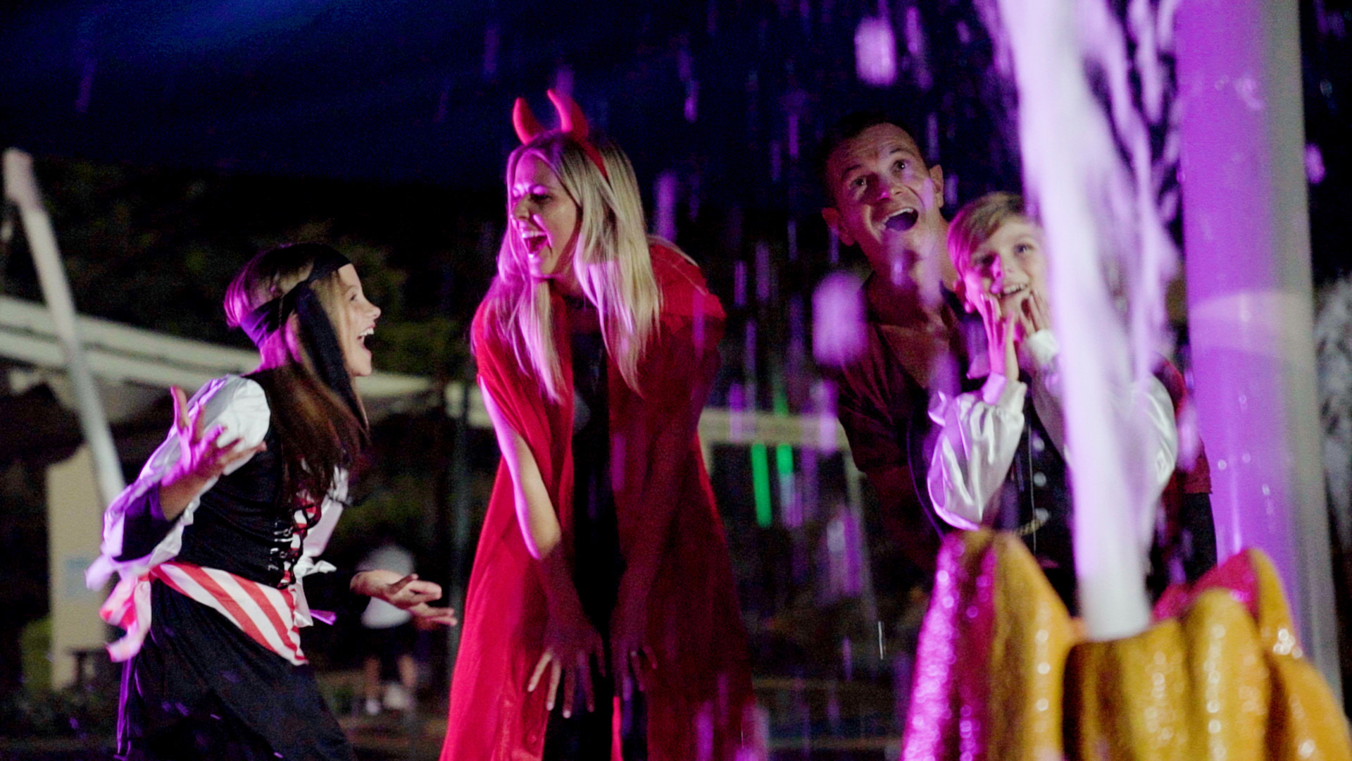 Four people in costumes—two adults and two children—laugh and play near a lit water fountain at night. One adult wears devil horns and a cape, while a child is dressed as a pirate.