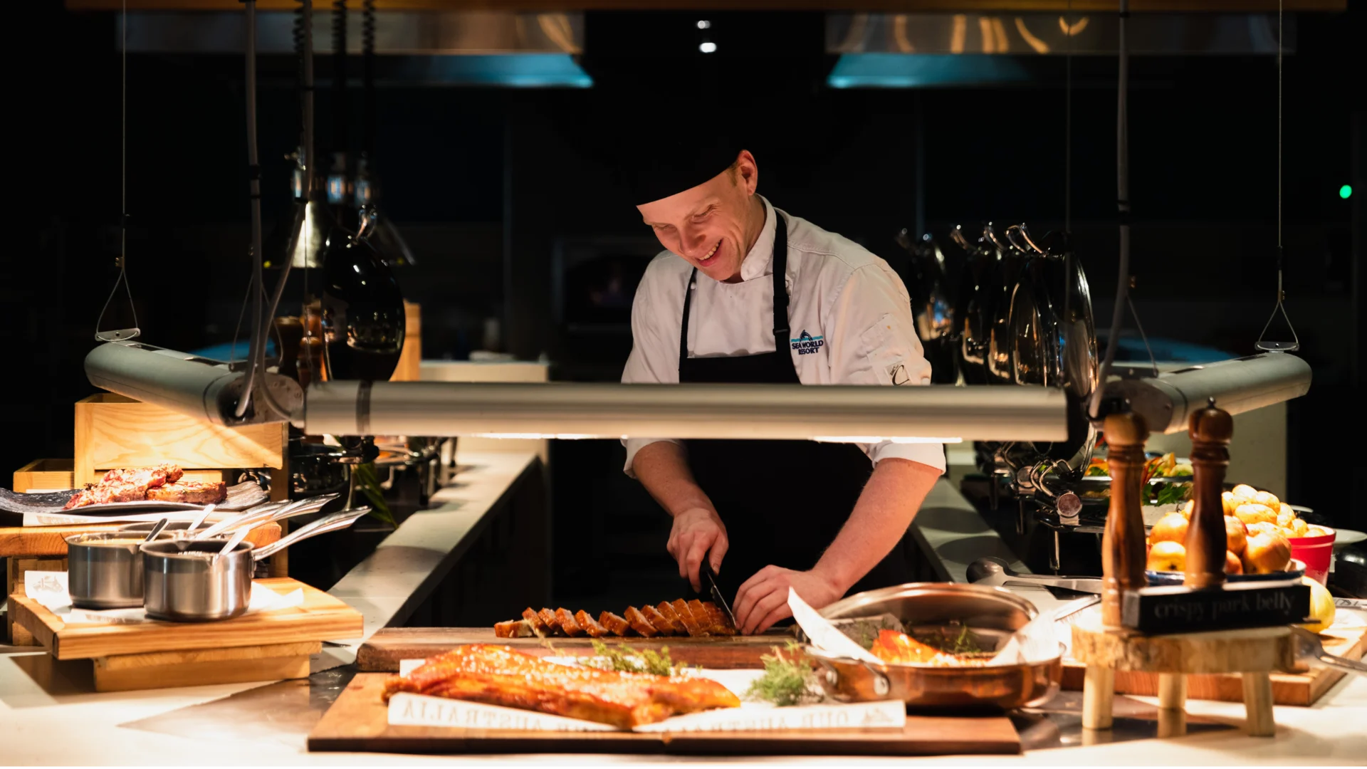 A smiling chef in a black hat and apron slices food on a wooden board in a well-lit restaurant kitchen, surrounded by various dishes and ingredients on the counter.