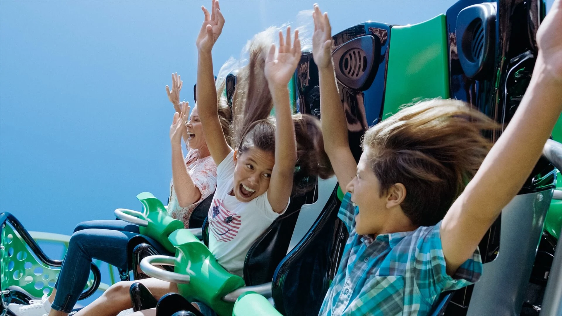 Three children with raised hands and excited expressions ride a green roller coaster on a clear, sunny day.