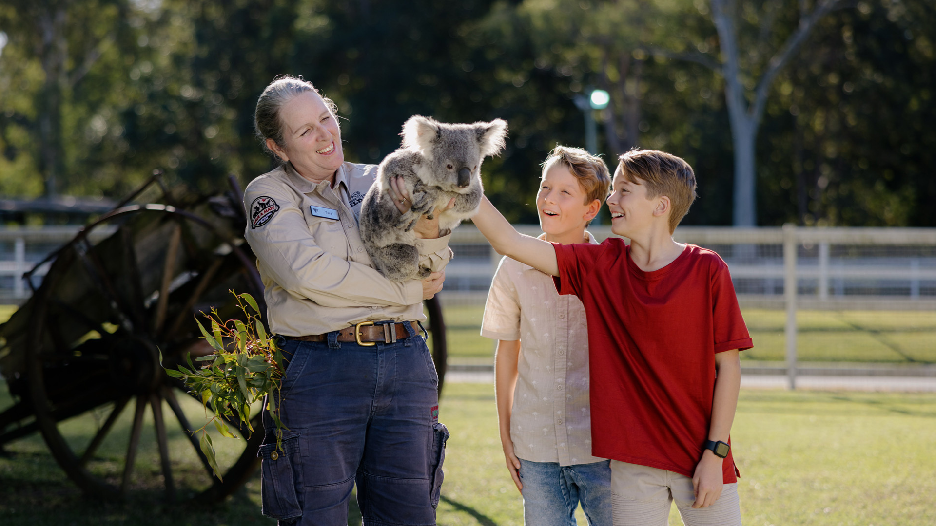 A park ranger holds a koala while two smiling children pet the animal outdoors near a wooden fence and old wagon wheel.
