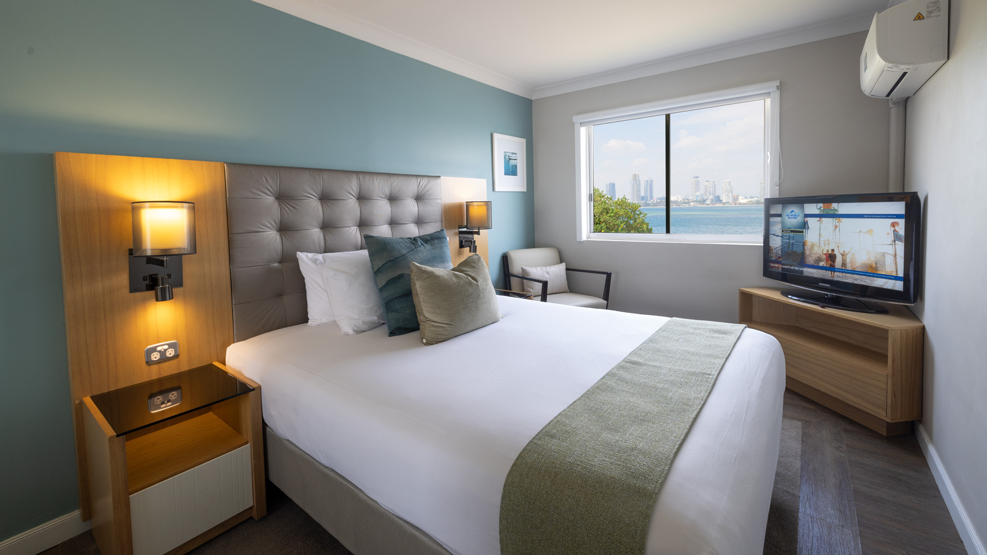 Modern hotel room with a large bed, green accent wall, and a window view of a city skyline over water. A TV and armchair are positioned near the window.