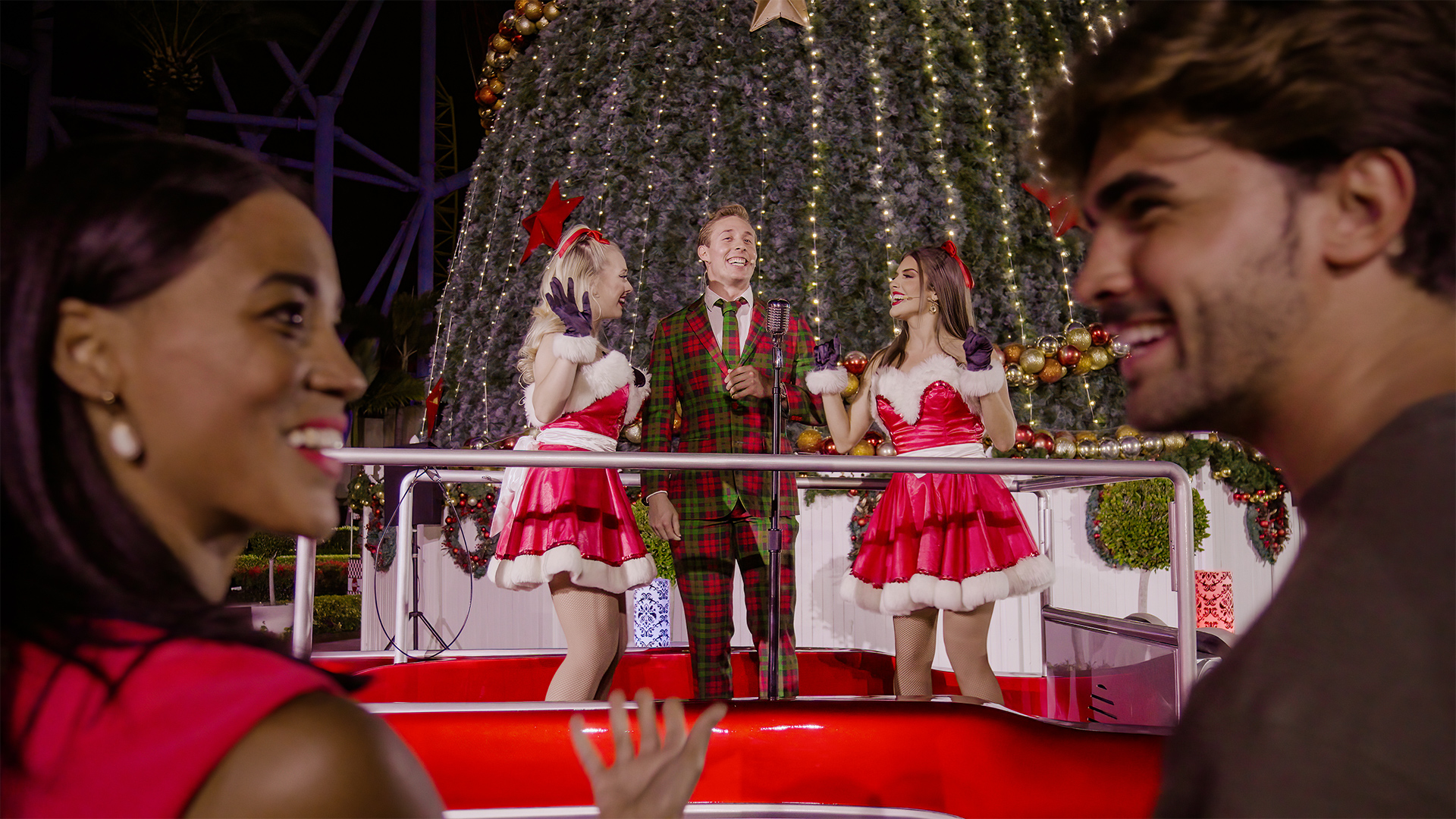 Two people smile in the foreground at a holiday event. Behind them, two women in red Santa dresses and a man in a plaid suit sing or perform in front of a large decorated Christmas tree.