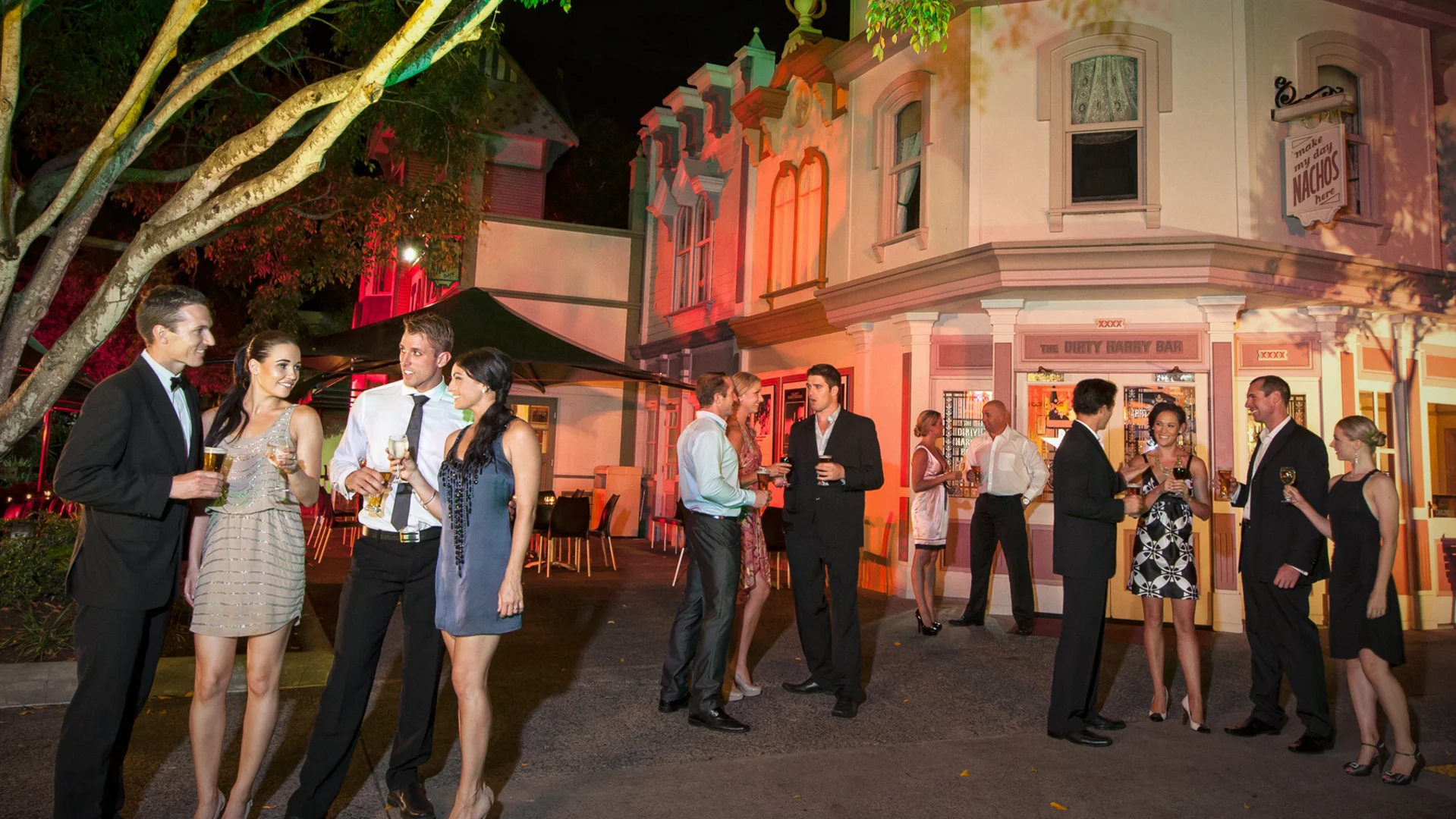 A group of people dressed in formal attire stand outside a brightly lit building at night, talking and holding drinks in a lively, festive atmosphere.