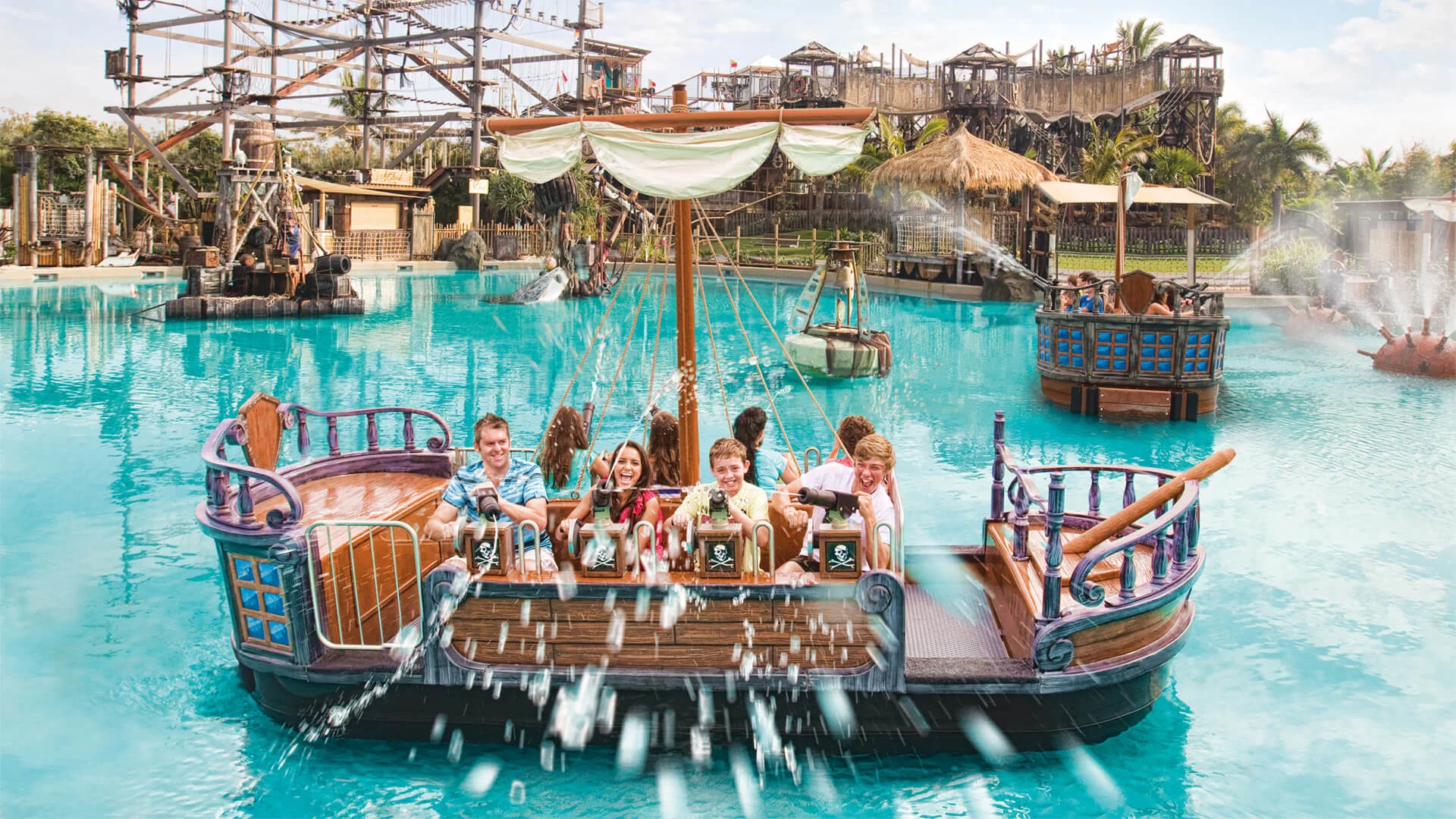 Children and adults ride a wooden-themed water boat attraction, splashing water in a large pool with a ropes course and wooden structures in the background.
