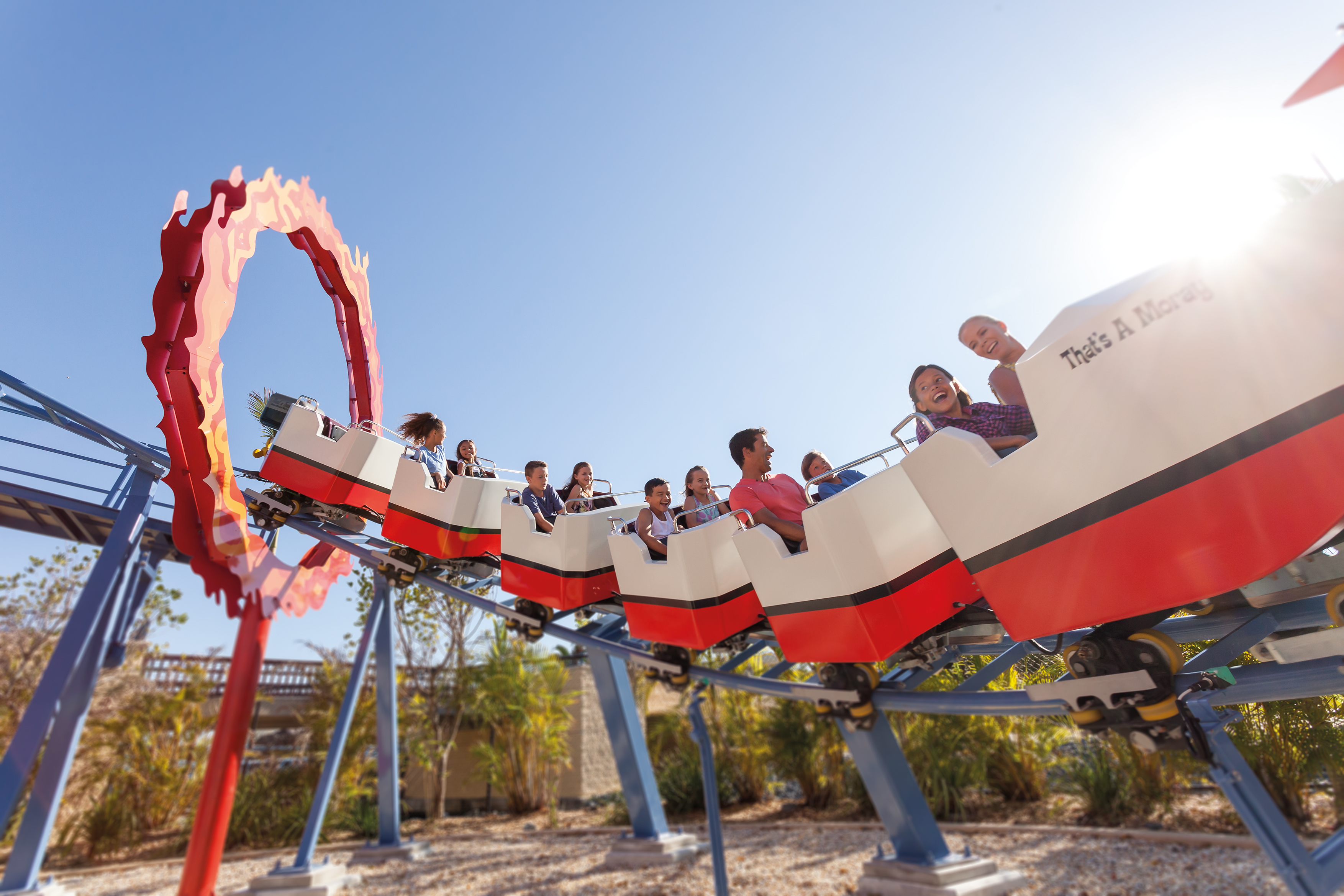 People ride a roller coaster passing through a red circular loop on a sunny day, with blue sky and trees in the background.