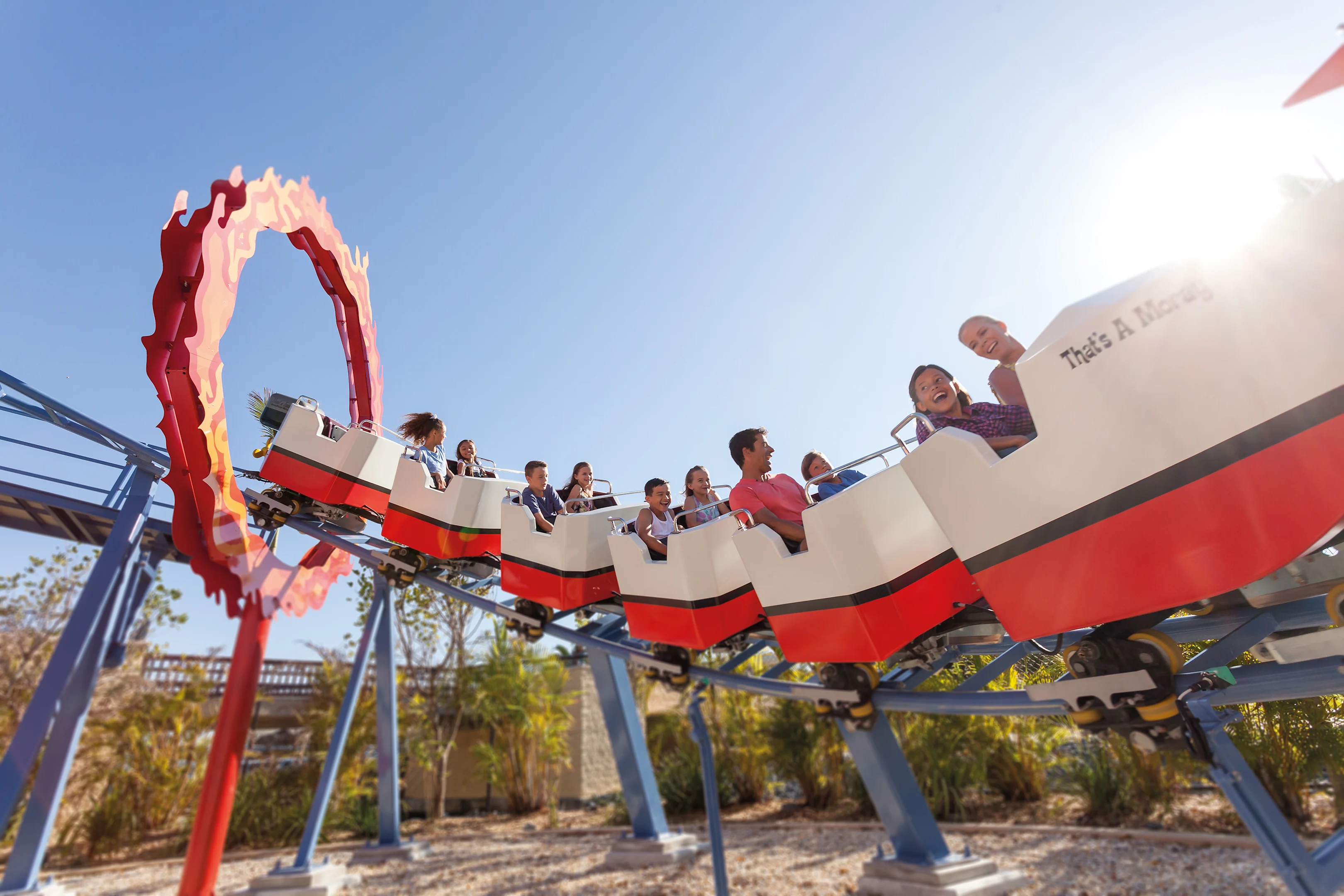 People ride a roller coaster passing through a red circular loop on a sunny day, with blue sky and trees in the background.