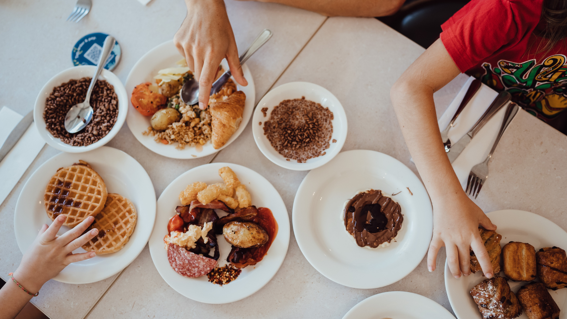 Overhead view of a table with various breakfast foods including waffles, cereal, pastries, fruit, and eggs. Three people are reaching for food, showing only their arms and hands.