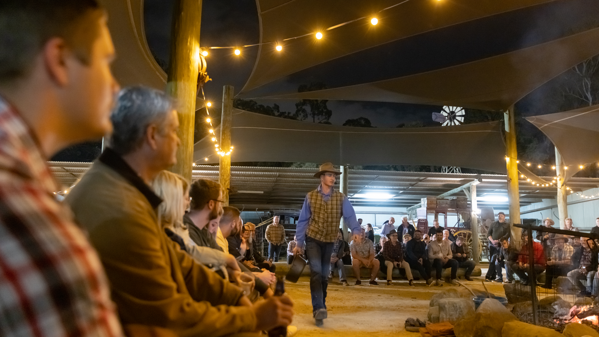 A group of people sit in a semicircle at night under string lights, watching a performer in a hat and plaid shirt walk in the center of an open, covered area.