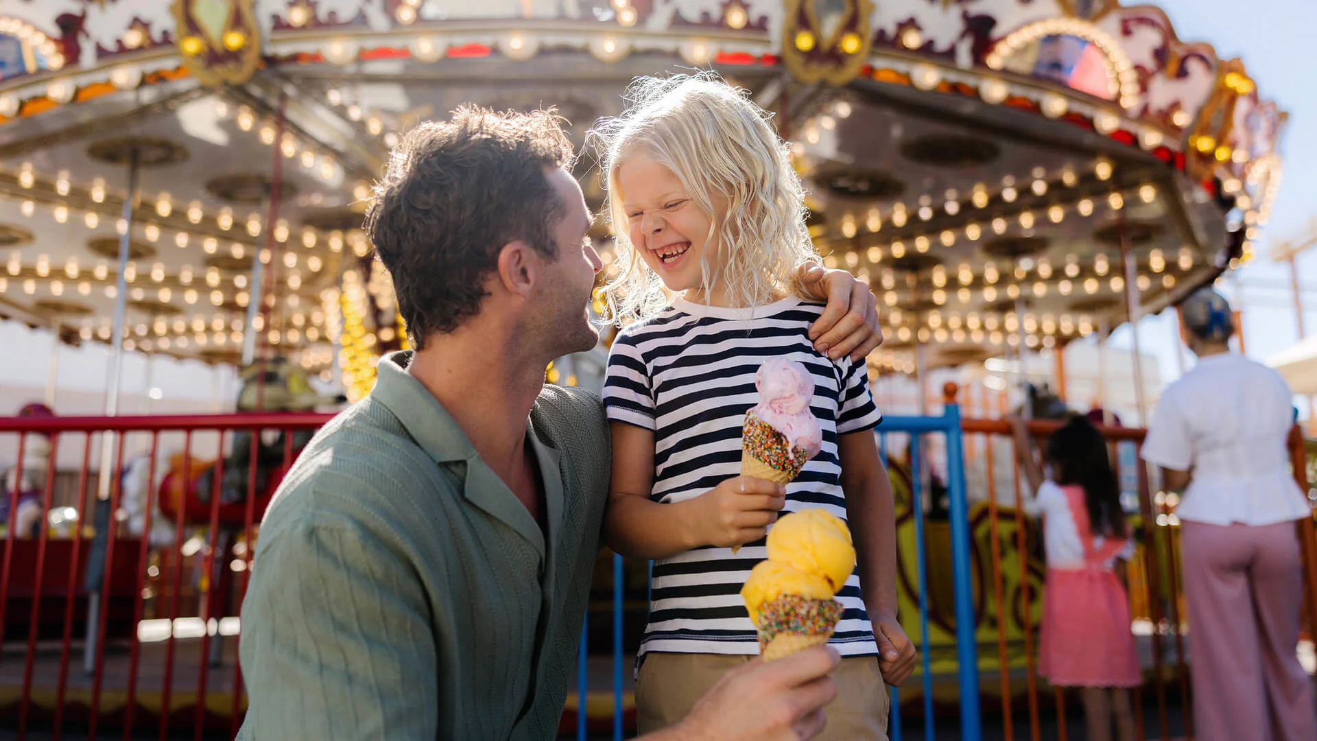 A man and a young girl smile at each other while holding ice cream cones in front of a brightly lit carousel at an amusement park. The girl wears a striped shirt and the atmosphere is joyful and festive.