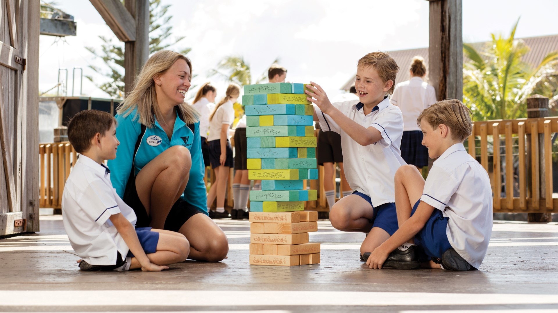 A woman and three boys in school uniforms play a large wooden block stacking game outdoors, smiling and laughing, with other children and trees visible in the background.