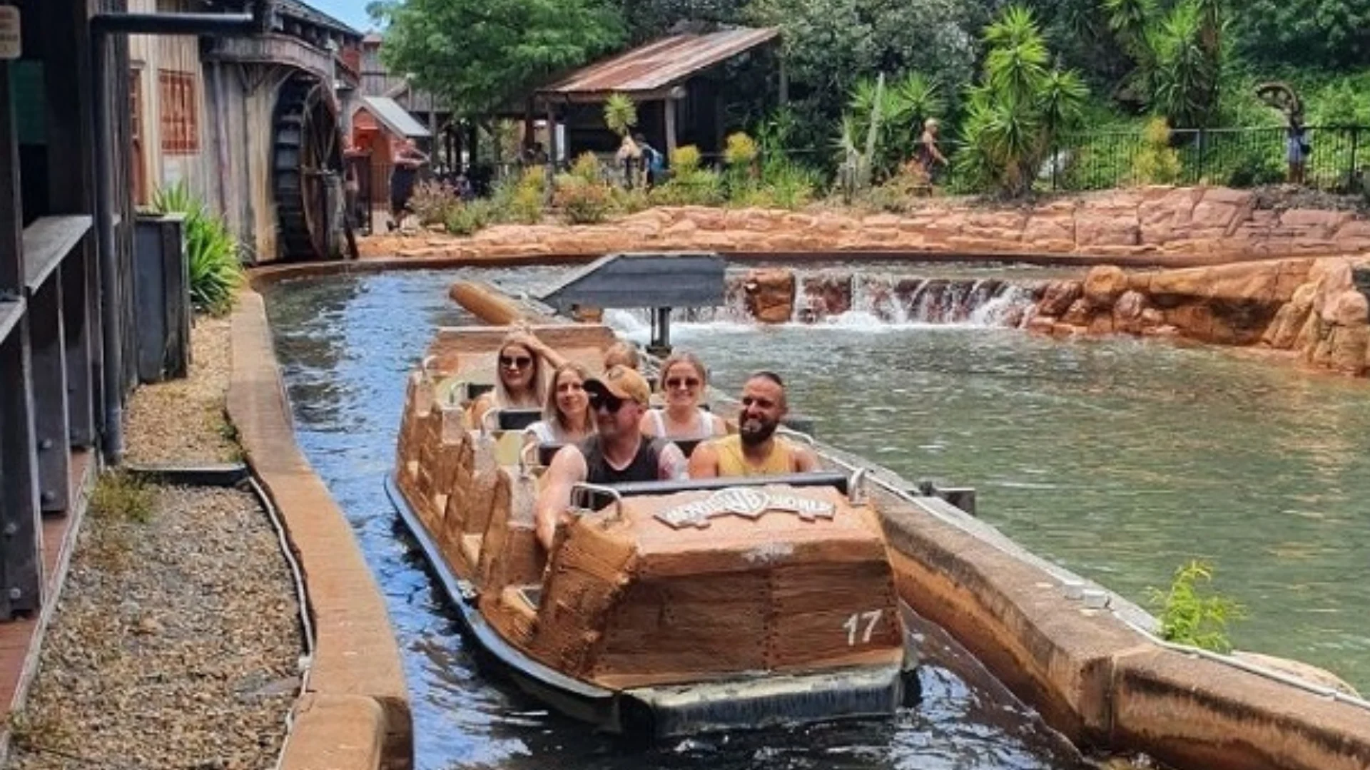 Five people ride a log flume boat on a water attraction, passing through a themed area with rocks, greenery, and buildings in the background. Everyone appears to be enjoying the experience.
