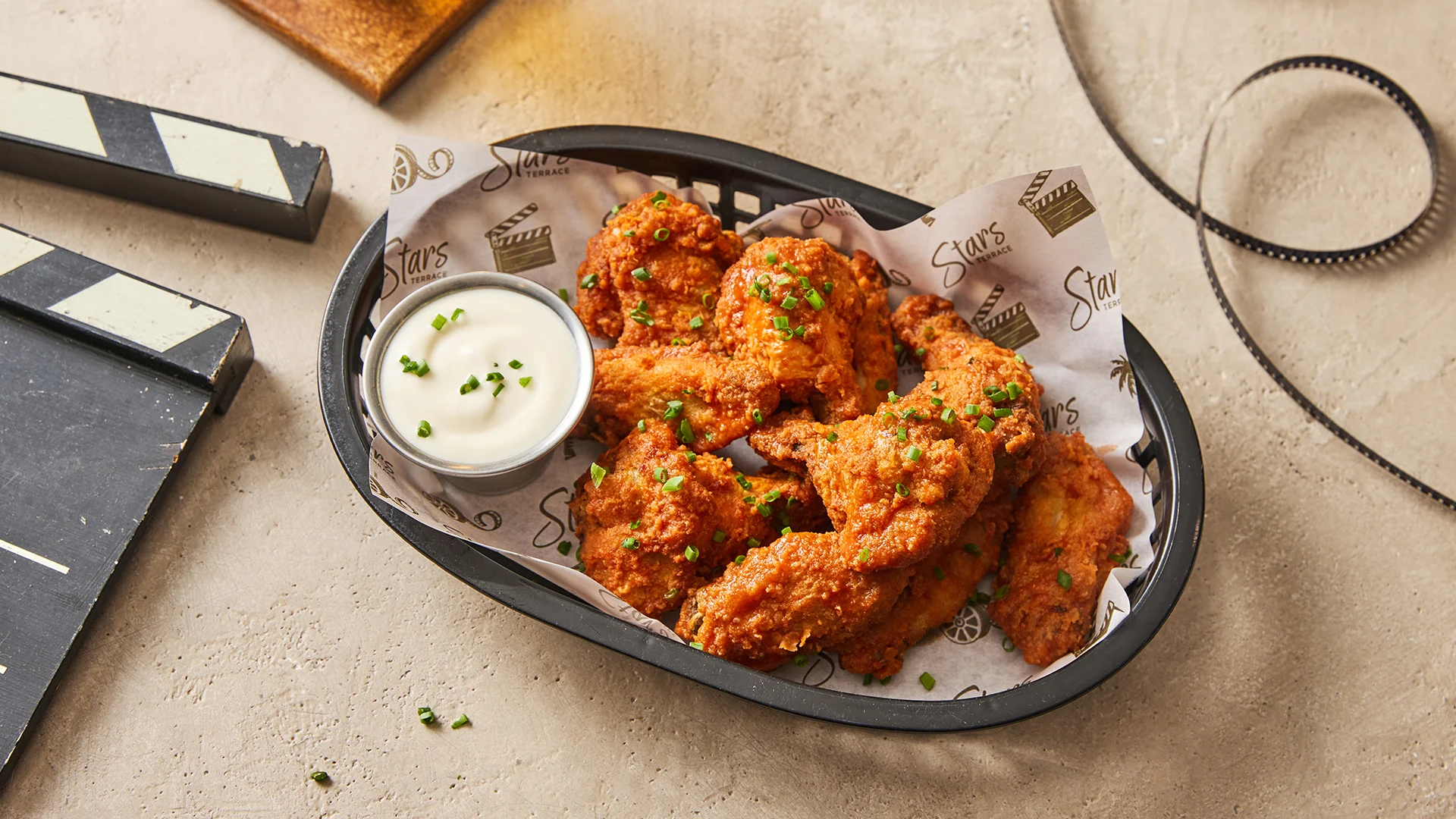 A black basket lined with paper holds crispy, saucy chicken wings garnished with chopped chives, served with a small cup of creamy dipping sauce on a light-colored surface.