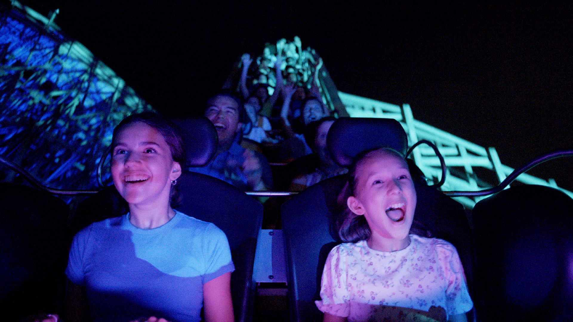 Two young girls in the front row of a roller coaster laugh and scream with excitement at night, while other riders behind them raise their hands and cheer under colorful lights.