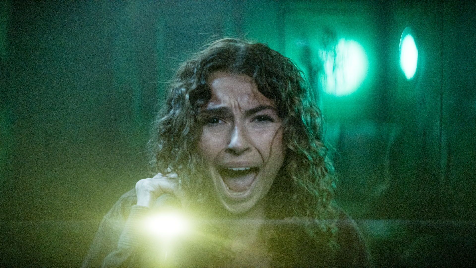 A young woman with curly hair screams in fear, holding a flashlight in a dimly lit, green-tinted room with blurred shapes and eerie lighting in the background.
