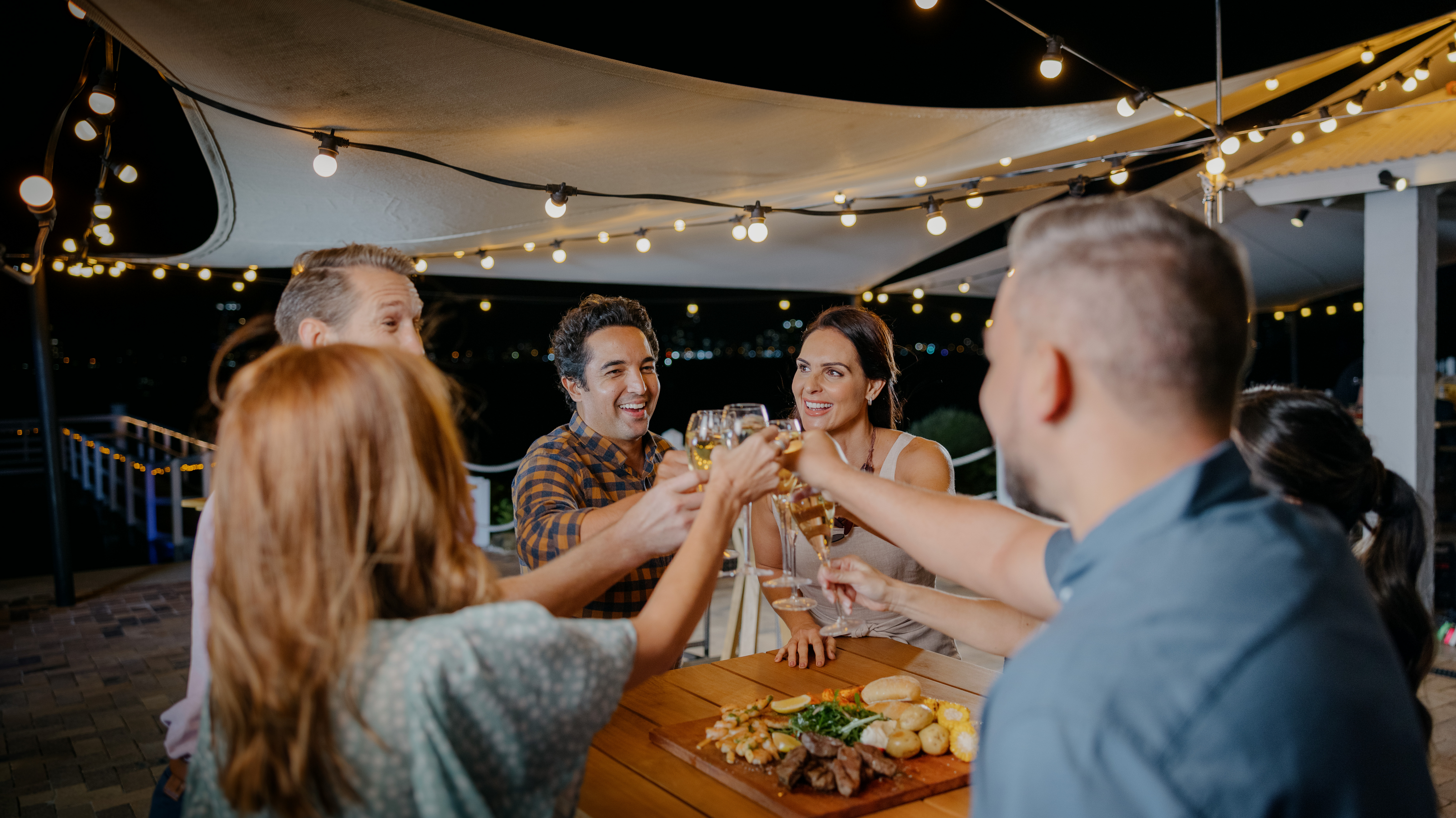 A group of people toast with drinks around a wooden table with food, under string lights at night.