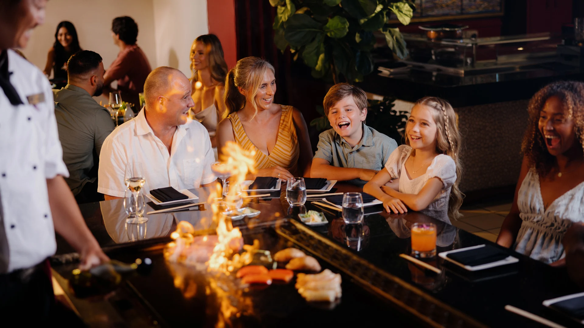 A group of people sit around a teppanyaki grill, watching the chef perform with flames at Hatsuhana Japanese Restaurant. Drinks and plates are on the table.