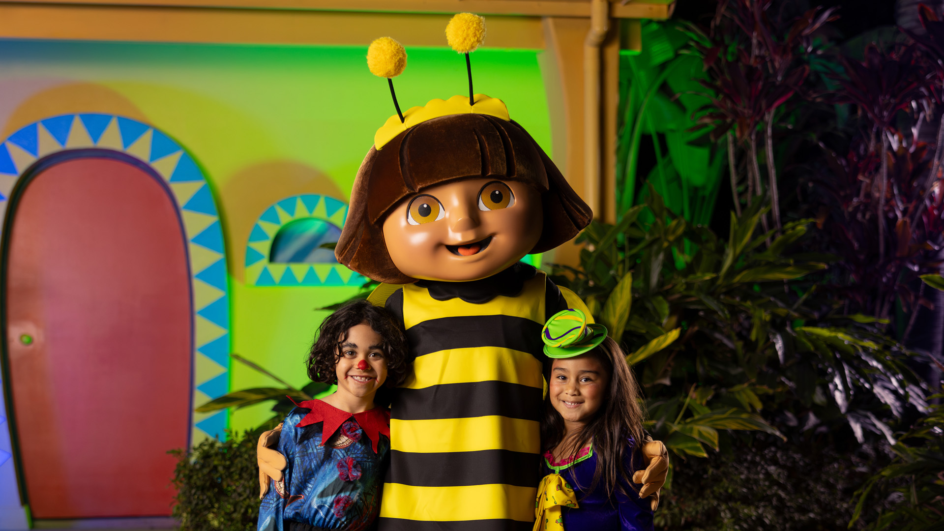 A person in a large bee costume with a big smiling face stands outside with two children in colorful costumes, all smiling and posing for the camera in front of a whimsical, colorful house.