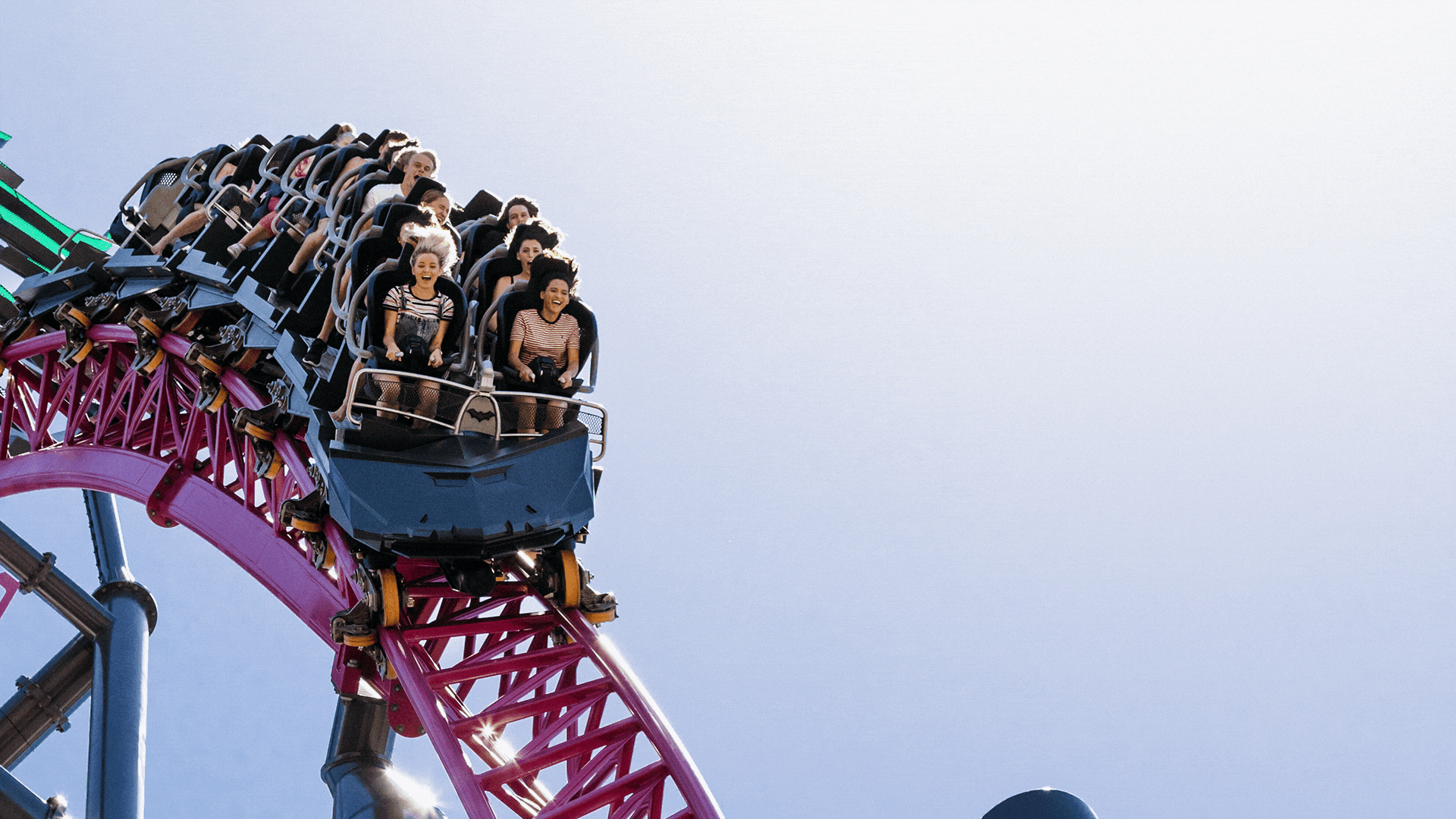 Several people are riding on a roller coaster, descending down a steep drop against a clear sky. The roller coaster track is painted in vibrant colors.