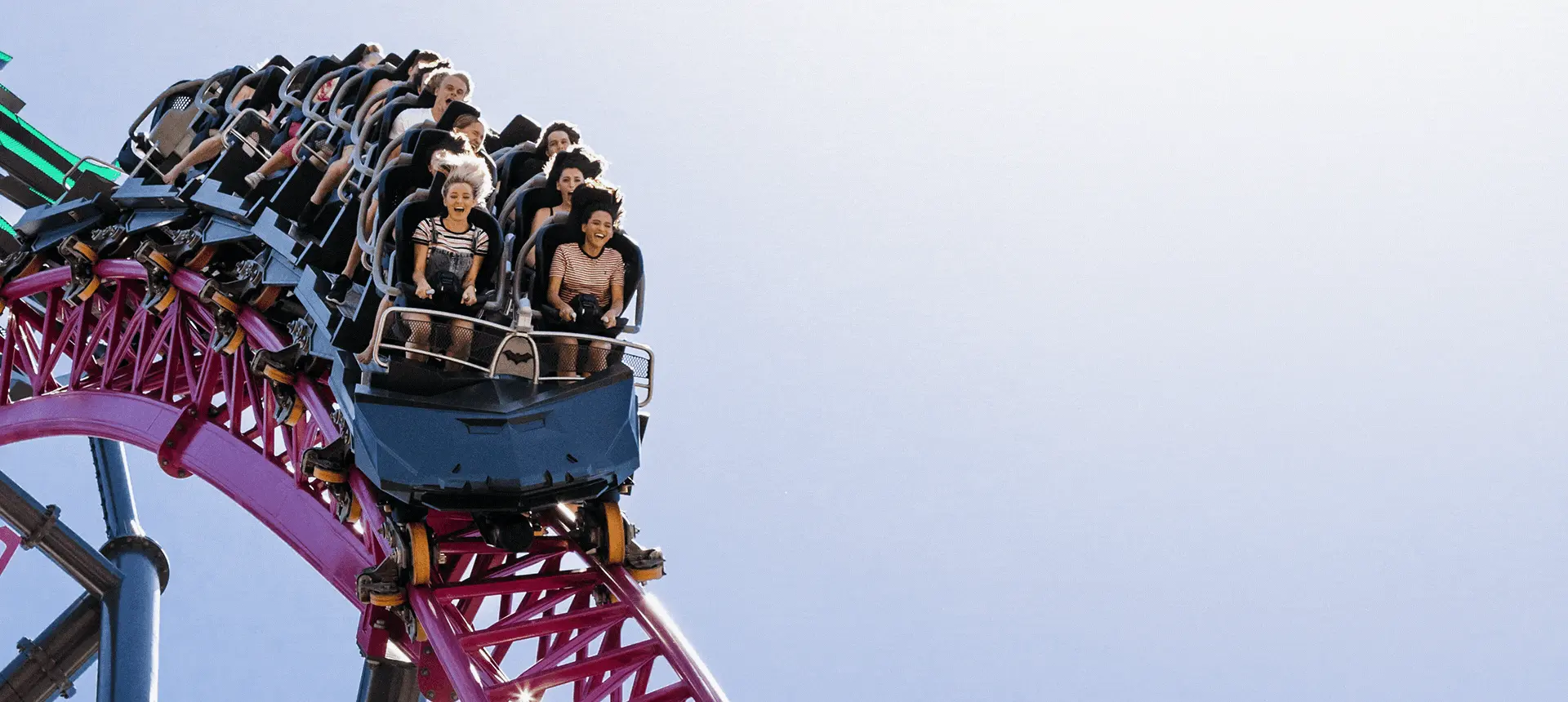 Several people are riding on a roller coaster, descending down a steep drop against a clear sky. The roller coaster track is painted in vibrant colors.