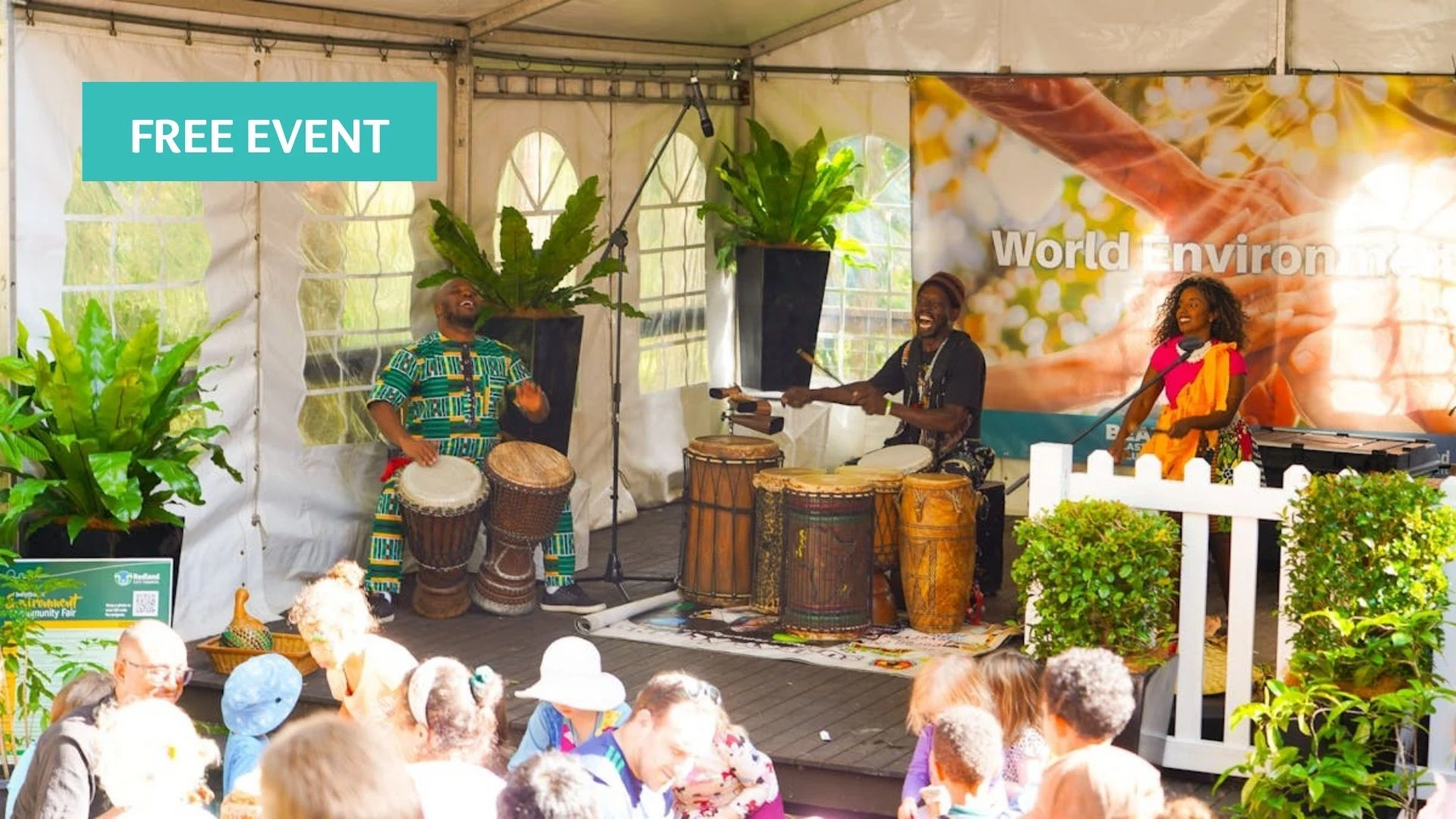 Three musicians in colorful clothing play drums and sing on a stage under a tent at a lively outdoor event, with green plants and a "FREE EVENT" sign visible. An audience watches in the foreground.