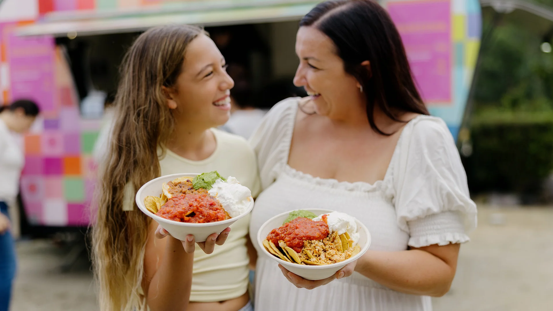 Two women smiling at each other while holding bowls of food topped with salsa, rice, chips, and sour cream, standing outdoors in front of a colorful food truck.
