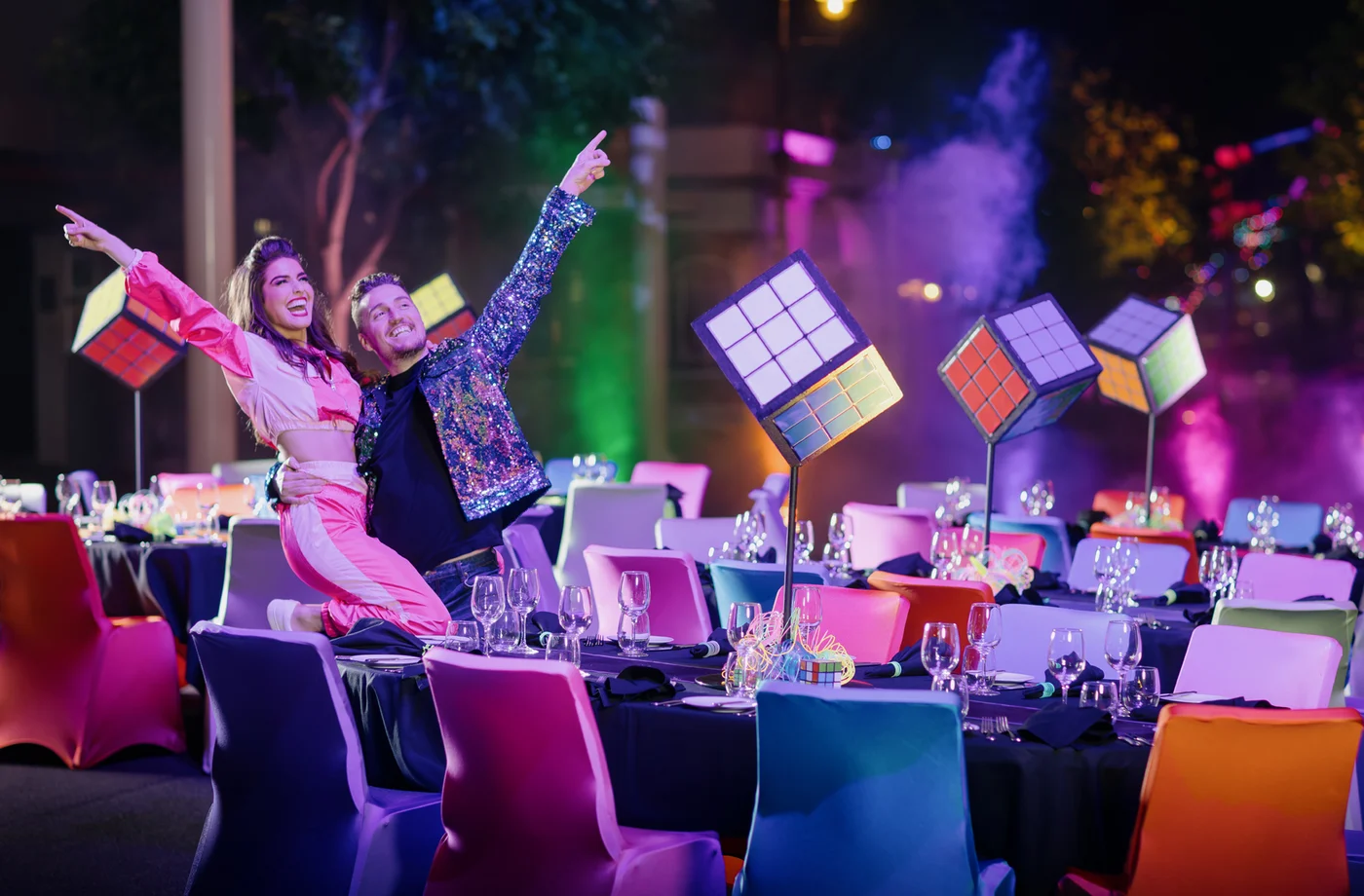 Two people in colorful outfits pose enthusiastically by a table at a vibrant, retro-themed event with Rubik’s cube centerpieces and multicolored chairs, surrounded by glowing lights and festive decor.
