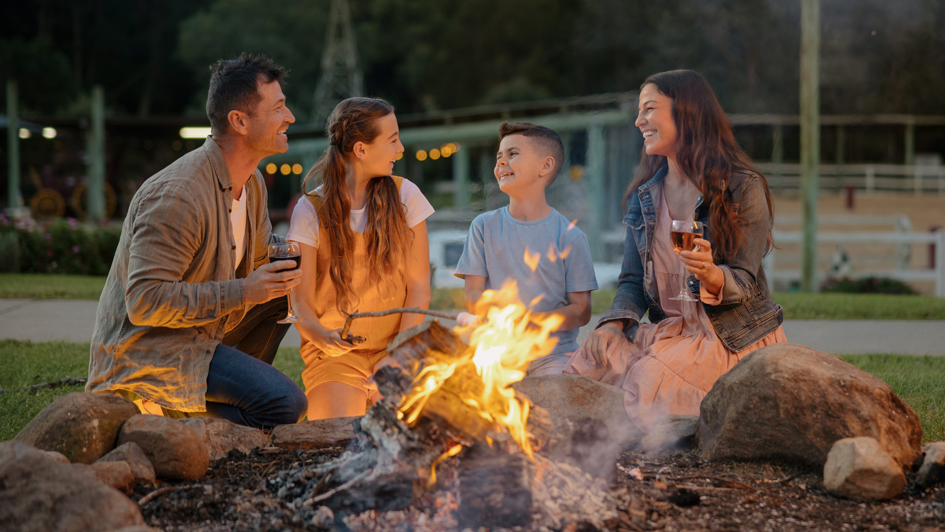 A family of four sits around a campfire outdoors in the evening, smiling and holding drinks.