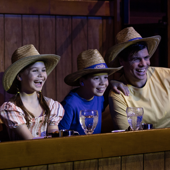 Three people wearing straw hats, two children and an adult, sit side by side at a wooden table with drinks, smiling and enjoying themselves in a rustic, warmly lit setting.