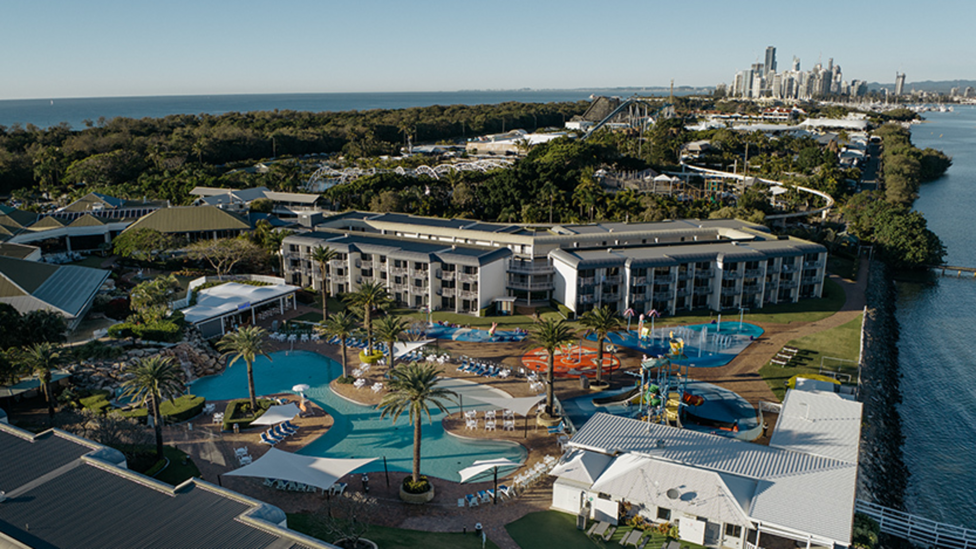 Aerial view of a large resort with swimming pools, palm trees, and colorful water play areas, surrounded by greenery and water, with a city skyline visible in the distance.