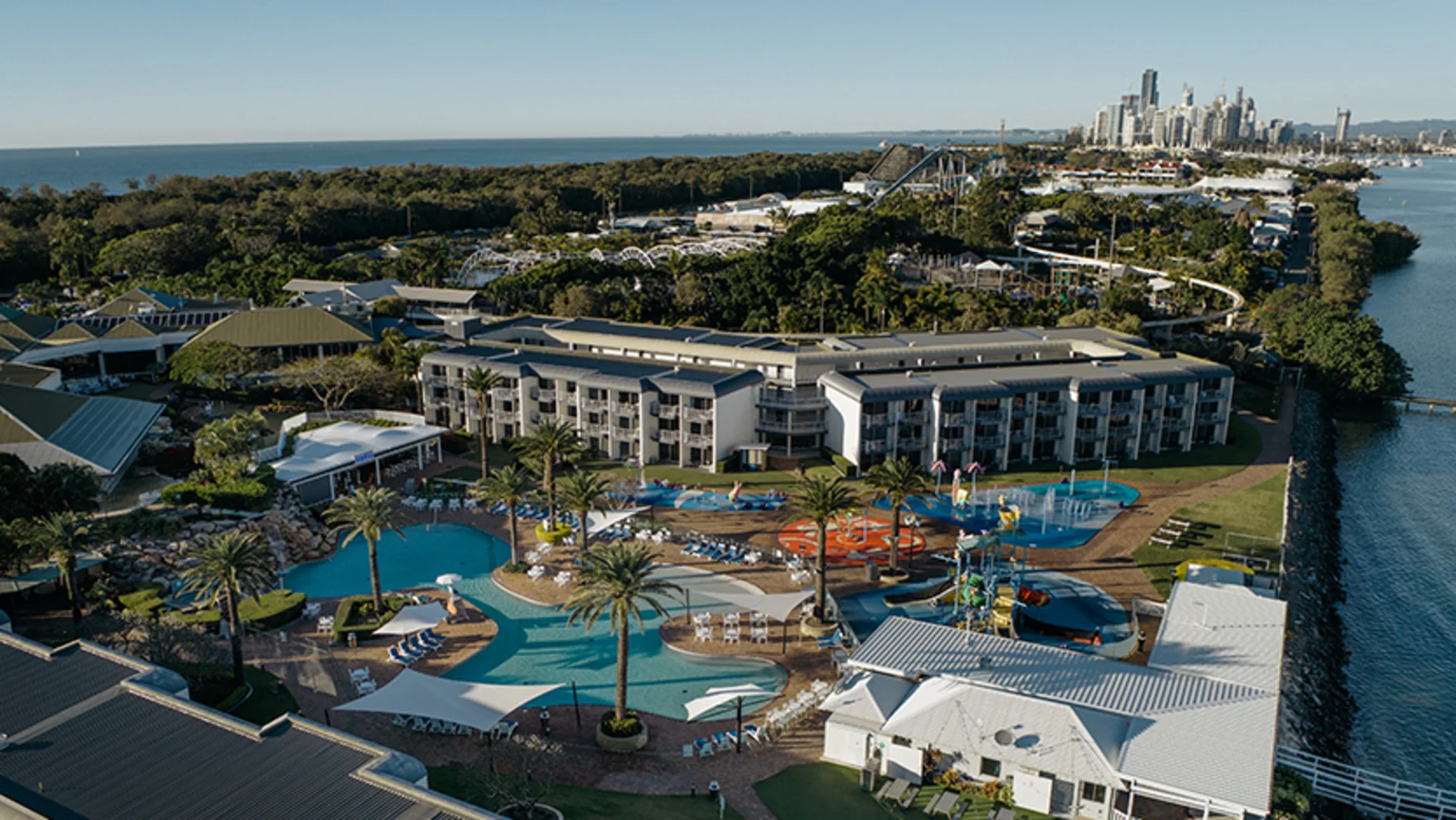 Aerial view of a large resort with swimming pools, palm trees, and colorful water play areas, surrounded by greenery and water, with a city skyline visible in the distance.