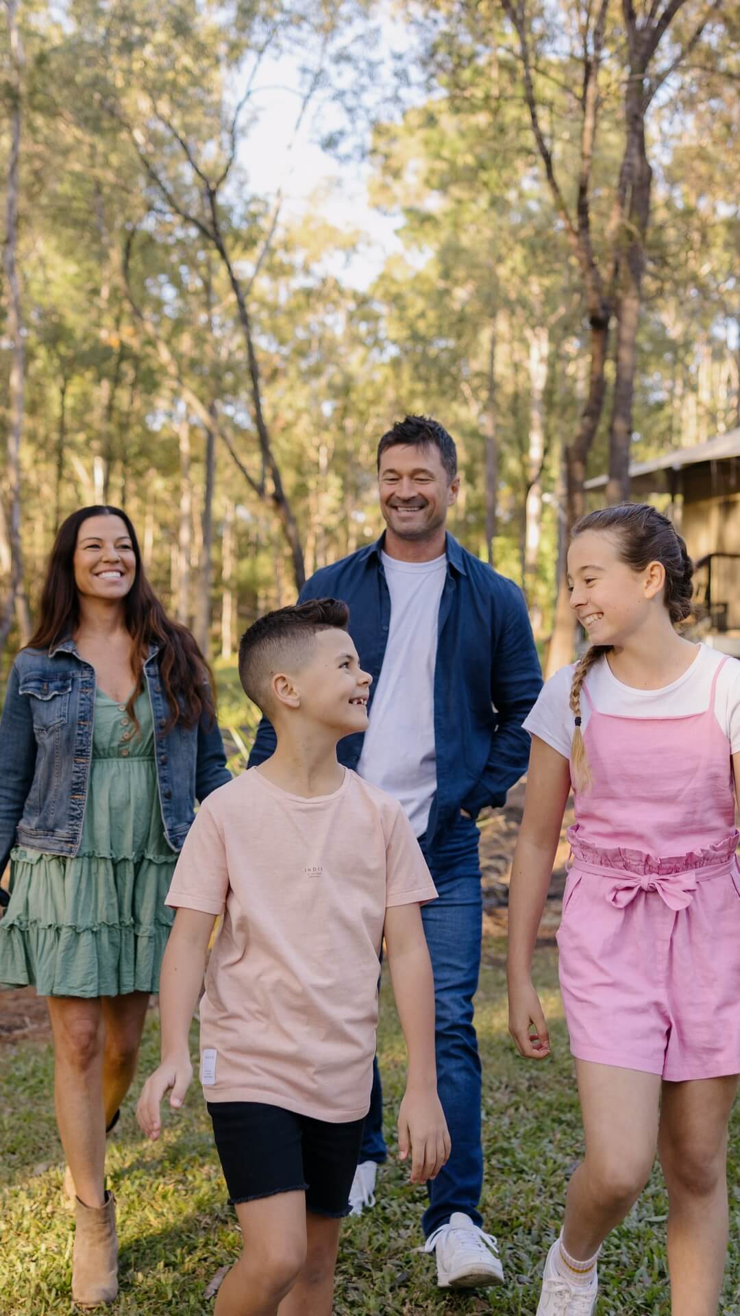 A family of four, including two children and two adults, walks together outdoors in a wooded area on a sunny day.