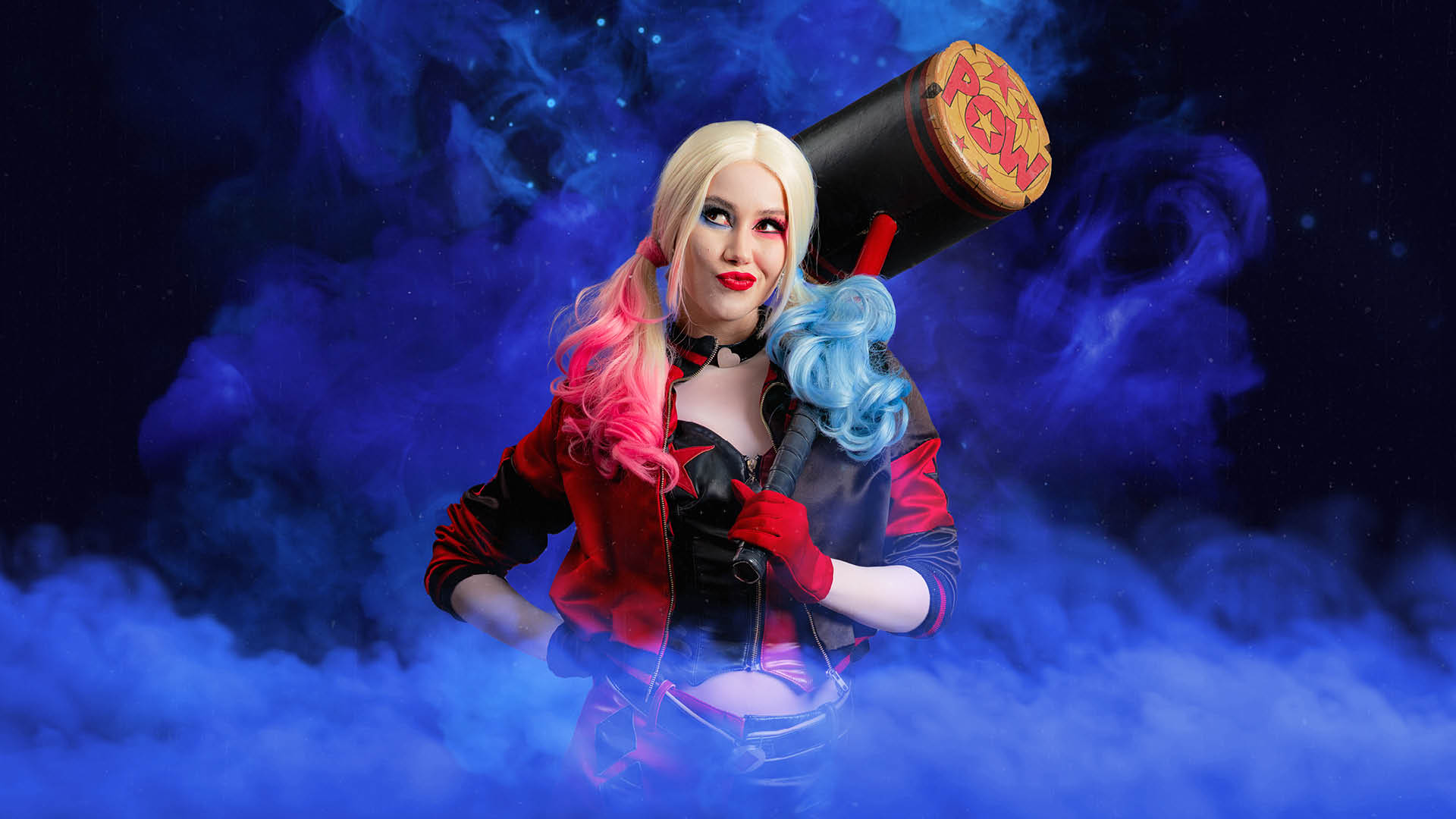 A woman dressed as Harley Quinn with blonde hair dyed pink and blue at the ends, wearing a red and blue costume, holds a large mallet over her shoulder, standing in front of smoky blue and purple background.