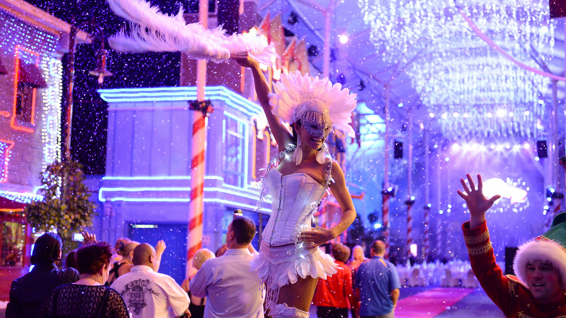 A performer in a white feathered costume entertains a crowd under bright, colorful lights at a festive, snowy indoor event. People watch, some waving, surrounded by holiday decorations.