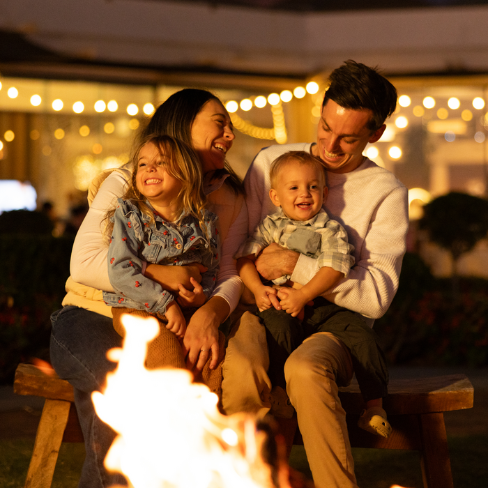 A happy family of four sits together on a bench by a campfire at night, smiling and laughing. String lights and a building are softly lit in the background, creating a warm, festive atmosphere.