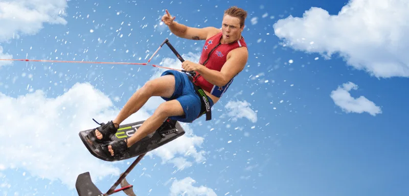 A man wearing a life vest and shorts is wakeboarding in mid-air against a blue sky with clouds, holding onto a tow rope.