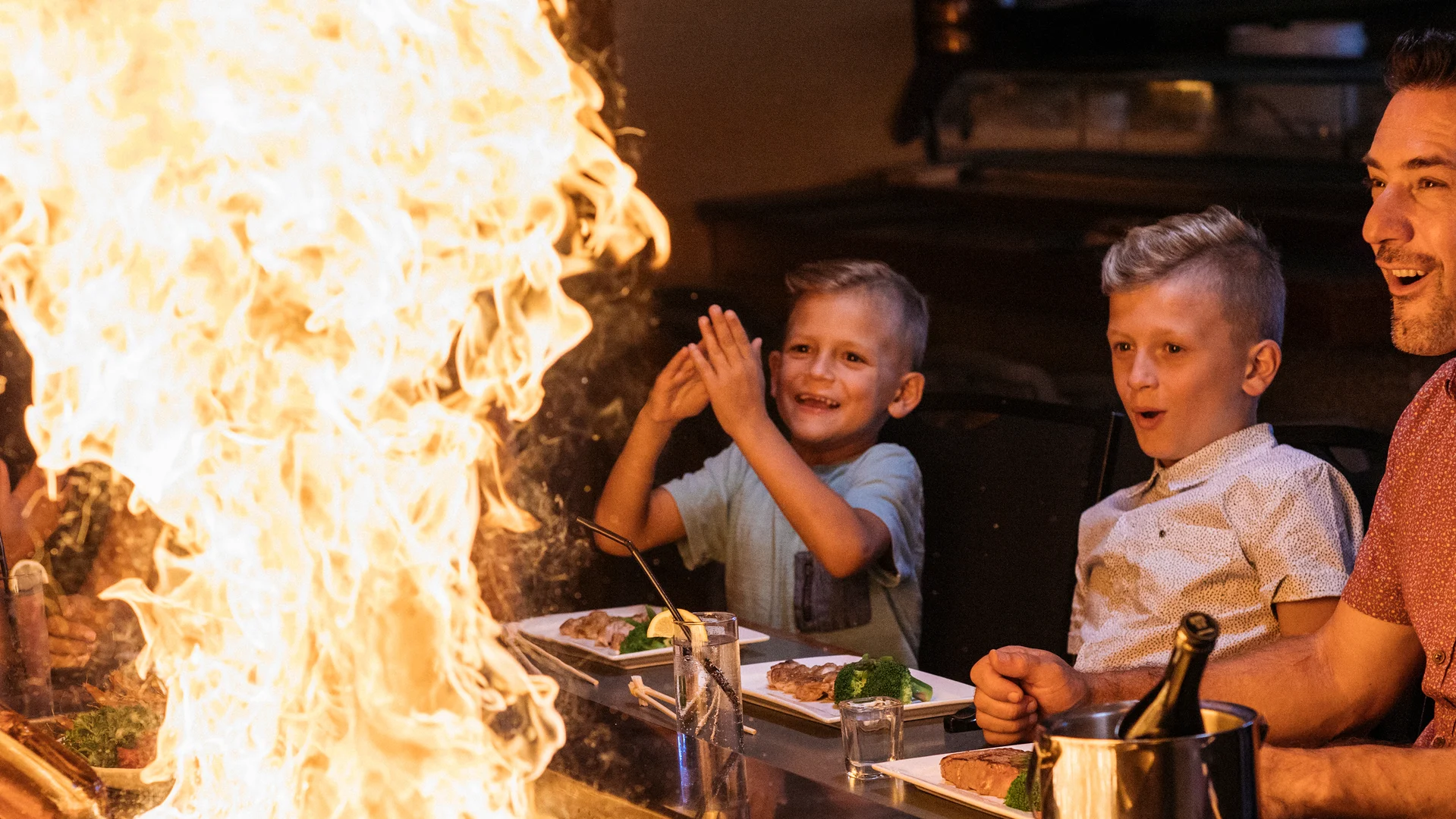 A chef creates a tall burst of flames on a hibachi grill while smiling children and an adult watch with excitement and surprise at a restaurant table.