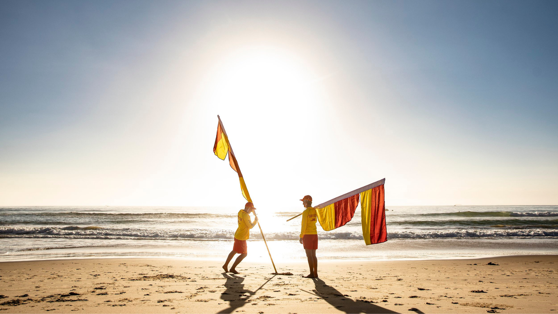 Two lifeguards in yellow shirts adjust red and yellow flags on a sunny beach.