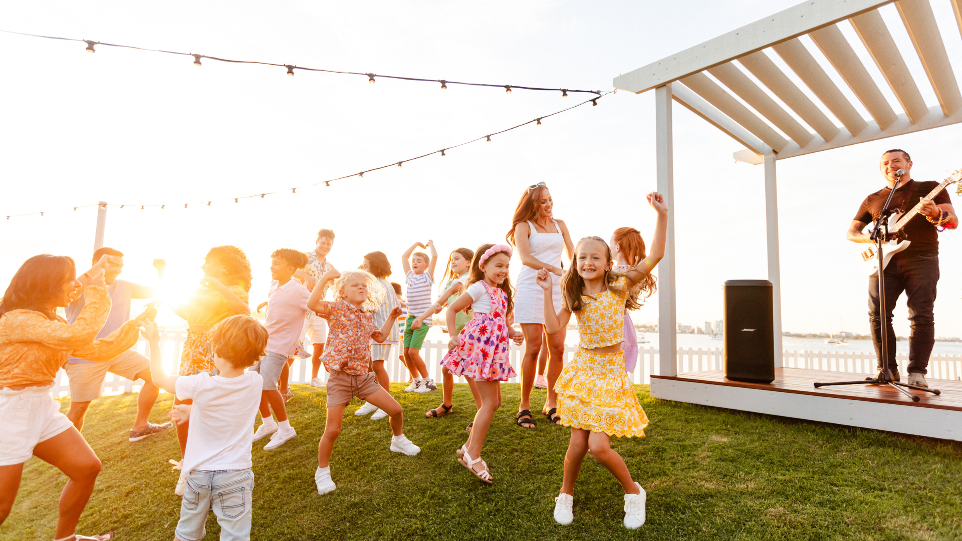 Children and adults dance and enjoy live music outdoors on a grassy lawn at sunset. A musician plays guitar on a small stage, while people laugh and celebrate under string lights.