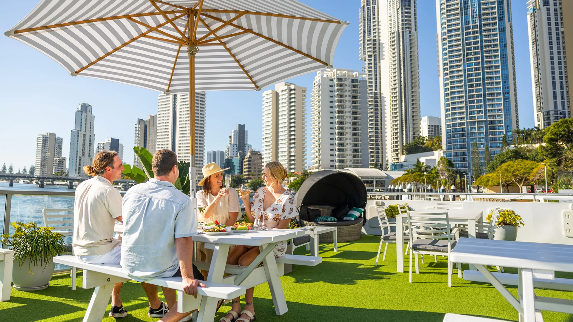 Four people sitting at an outdoor table on a grassy terrace by water, surrounded by tall buildings, under a large umbrella, on a sunny day.