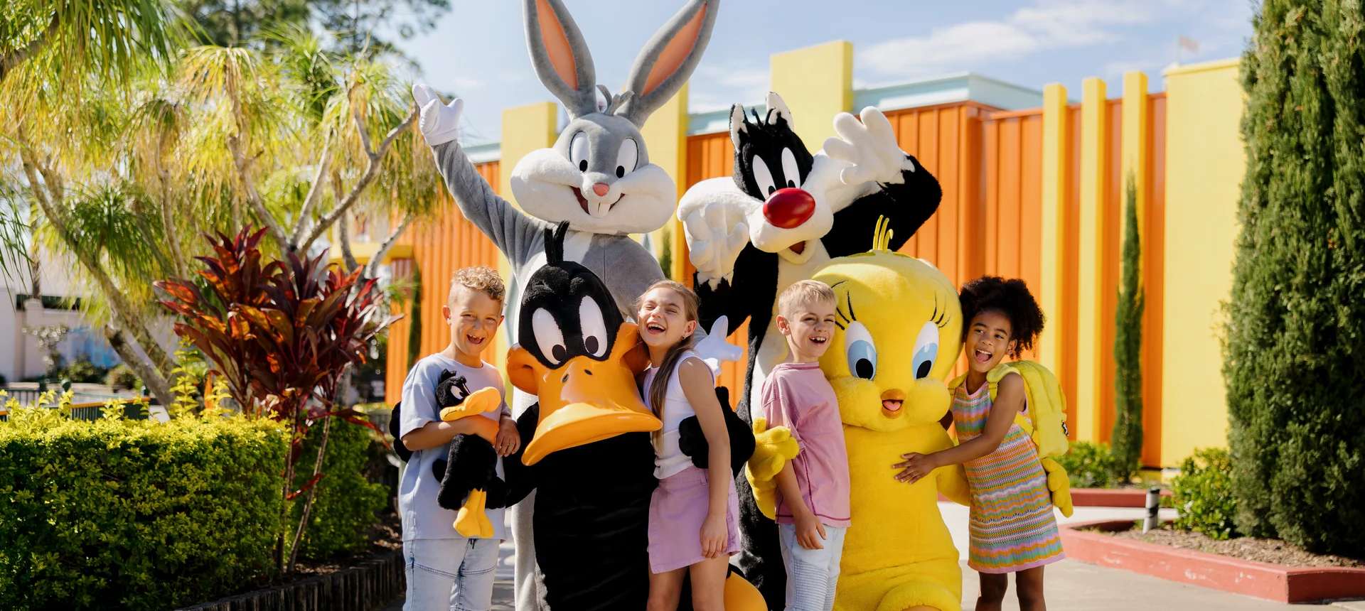 Four smiling children pose outdoors with people dressed in Bugs Bunny, Daffy Duck, Sylvester the Cat, and Tweety Bird costumes, surrounded by greenery and colorful buildings on a sunny day.