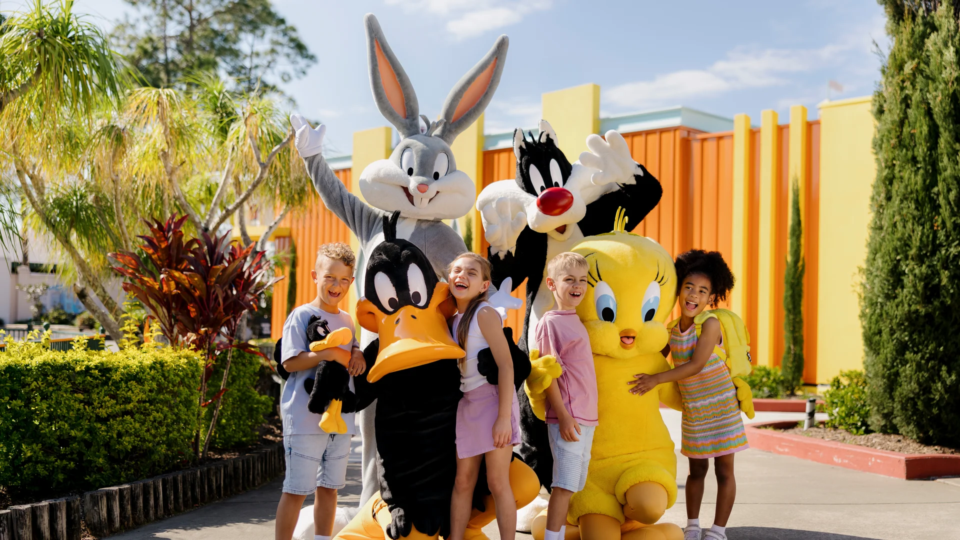 Four smiling children pose outdoors with people dressed in Bugs Bunny, Daffy Duck, Sylvester the Cat, and Tweety Bird costumes, surrounded by greenery and colorful buildings on a sunny day.