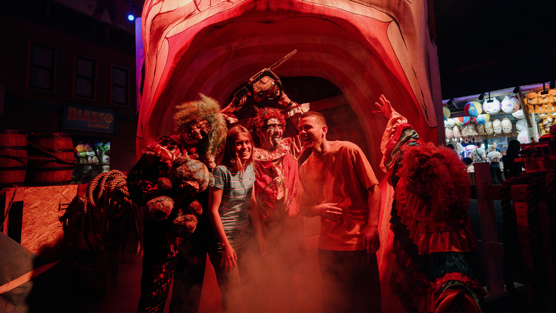 Two smiling people pose with actors in creepy monster costumes under a giant open mouth prop at a spooky, foggy amusement park attraction with colorful lights and booths in the background.