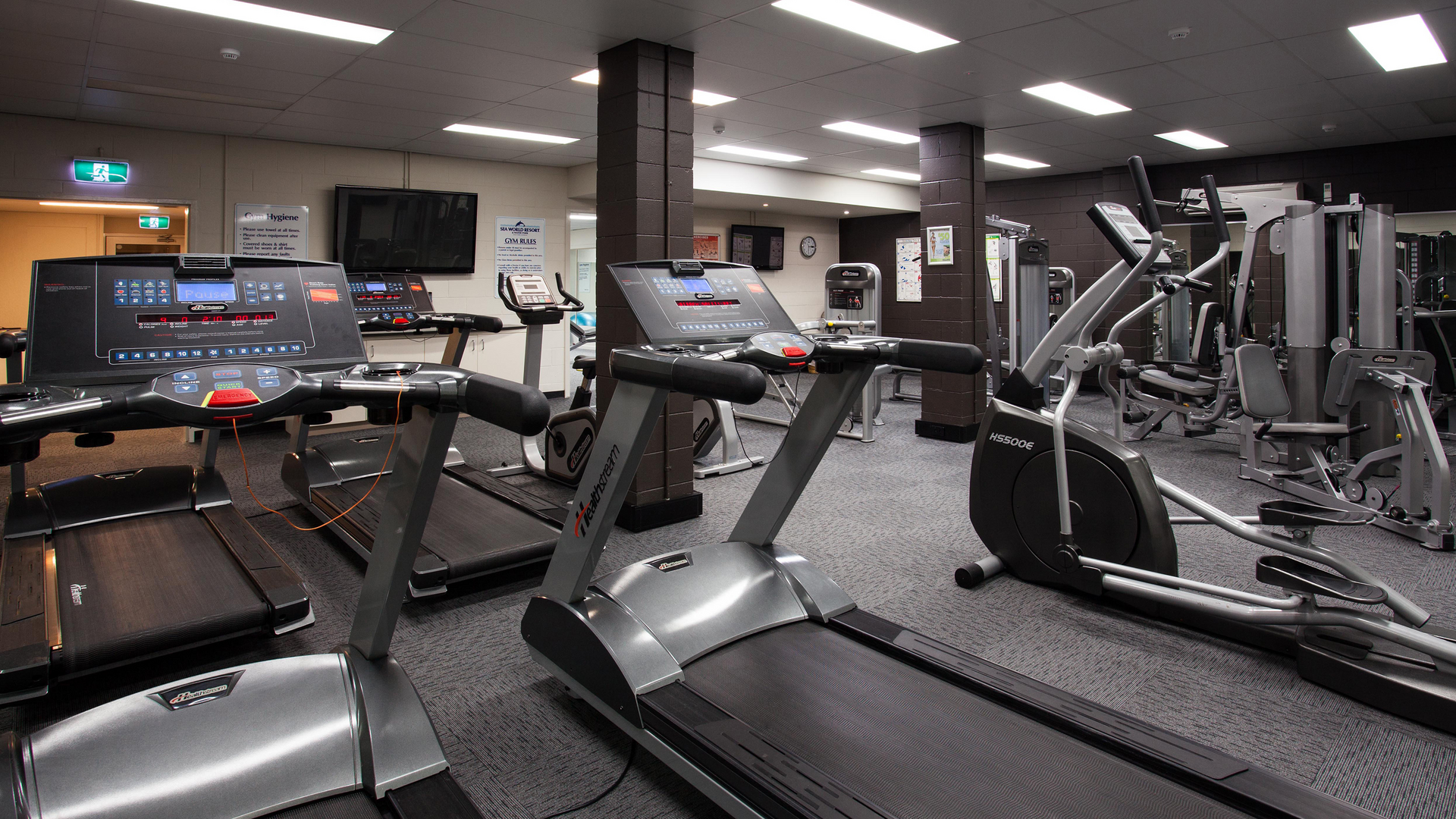 A modern gym with several treadmills, elliptical machines, and various strength-training equipment. The room has grey carpeting, bright lighting, and mirrors on the walls, creating a spacious and clean workout environment.